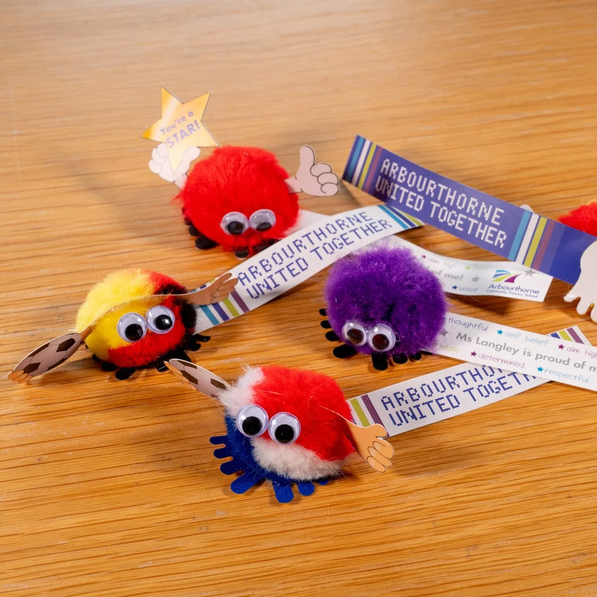 Colorful pom-pom spiders with googly eyes and paper arms holding 'Arbouthorne United Together' banners and star-shaped signs on a wooden surface.