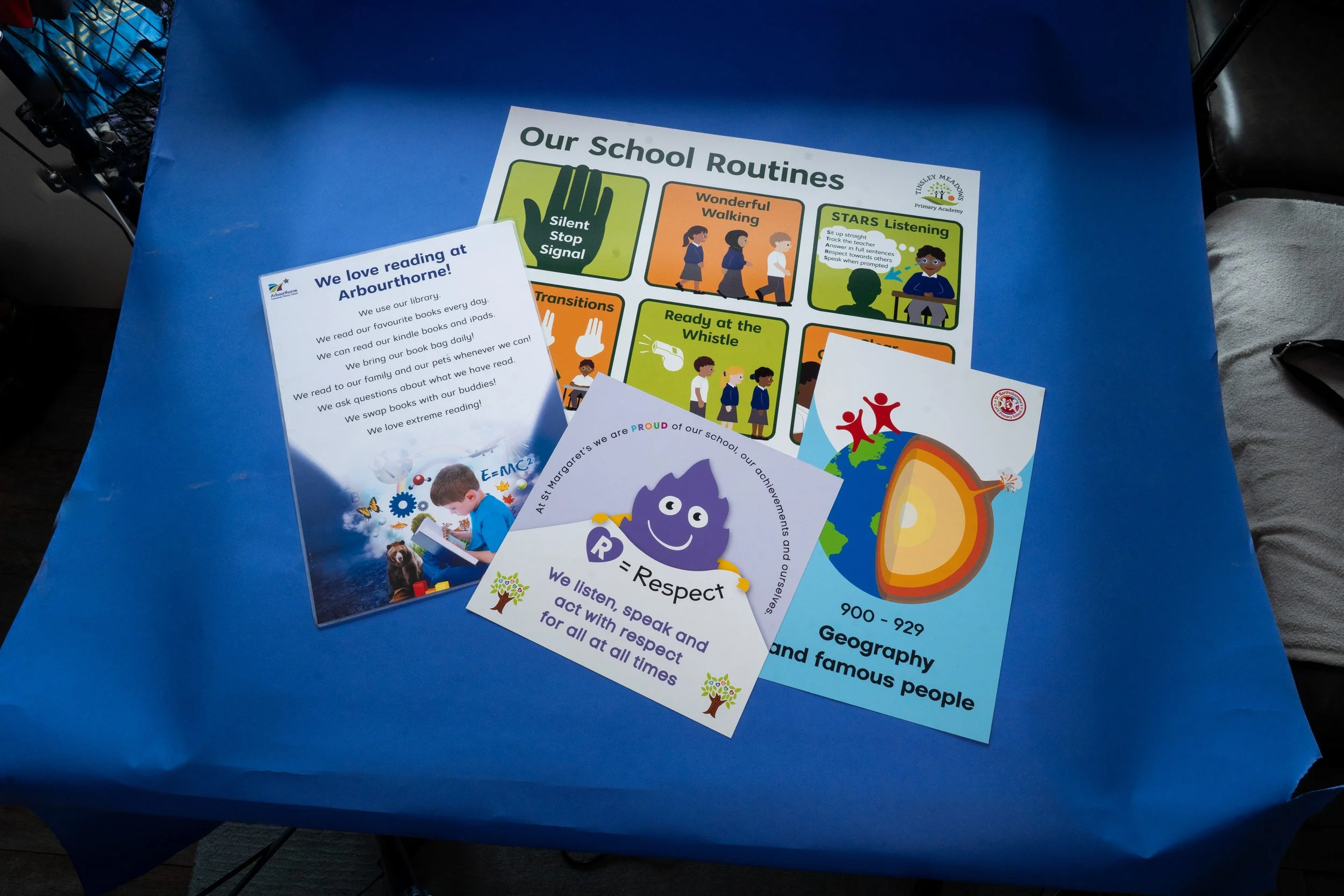 A blue table with various colorful printed flyers and posters about school routines and activities, including topics like walking, transitions, listening skills, and reading at Arbourthorne
