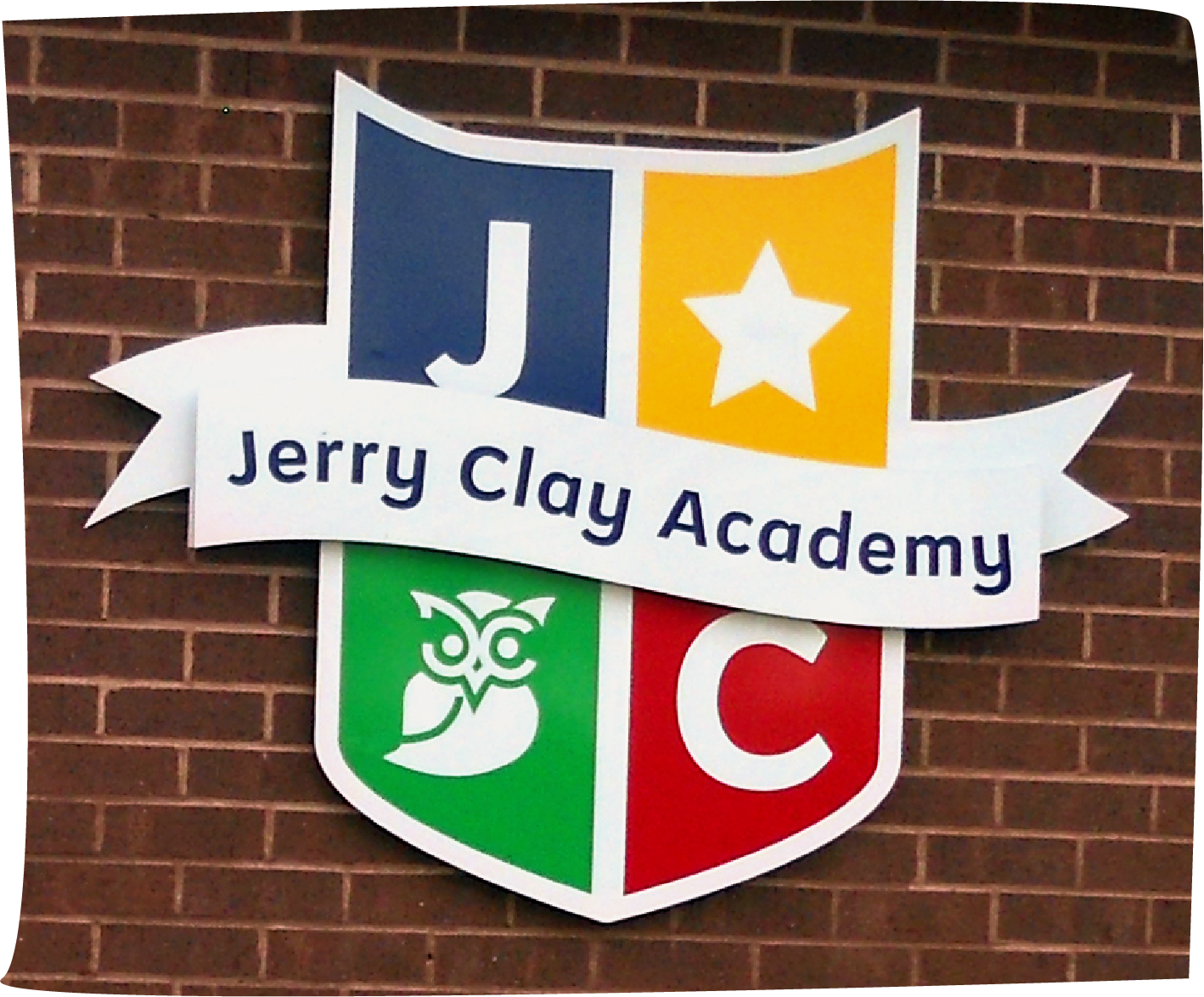 Jerry Clay Academy's shield-shaped logo on the wall outside the school. The shield logo is formed from 4 different colour quadrants, featuring the letters J & C, an owl and a star. The school’s name is emblazoned across an offset white ribbon banner