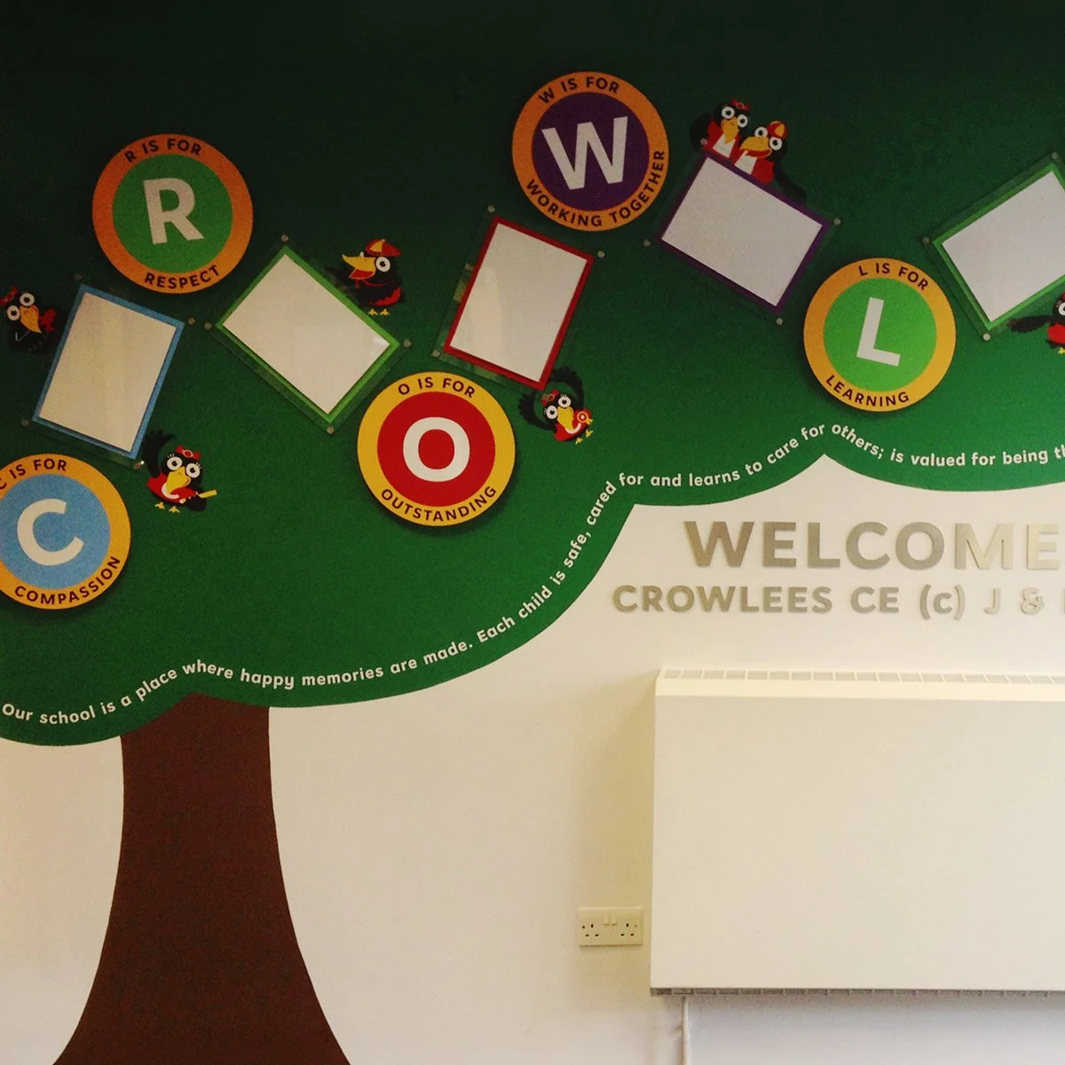 Decorative school wall display with colorful circles representing characters and values, a tree trunk graphic, and welcome message for children at Crowlees CE school.