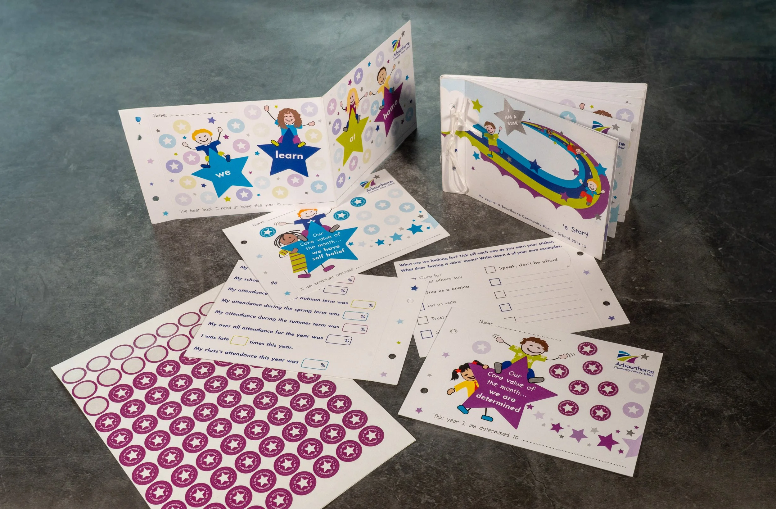 A collection of colorful student progress reports and stickers on a gray surface. The reports feature cartoon illustrations of children with motivational messages, and the stickers are circular with a star design.