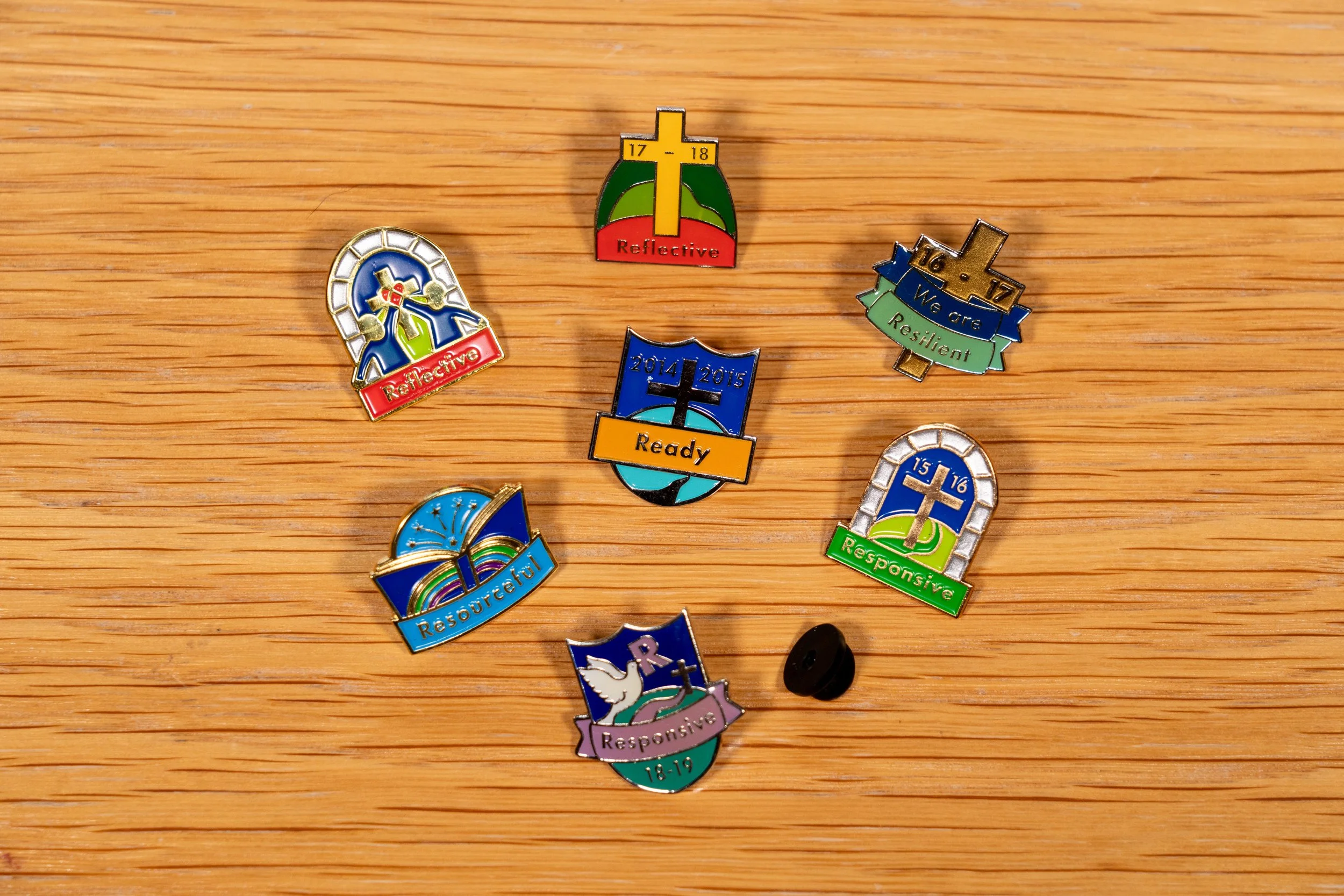 Collection of colorful enamel pins on a wooden surface, each with inspirational words like 'Reflective,' 'Resilient,' 'Resilient,' 'Responsible,' and 'Ready,' featuring various symbols and designs.