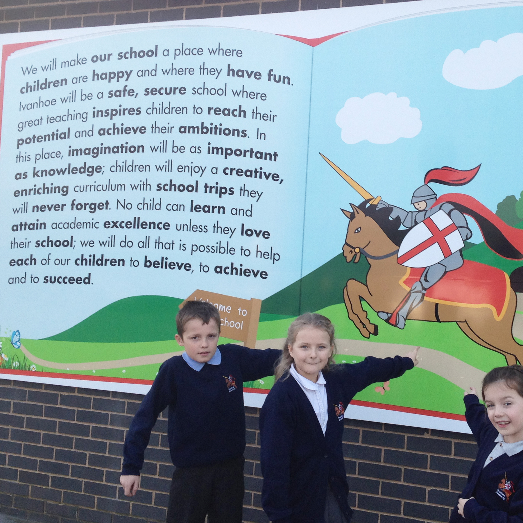 A group of school children standing in front of a colorful school sign with a cartoon knight riding a horse. The children are wearing school uniforms and pointing at the sign.