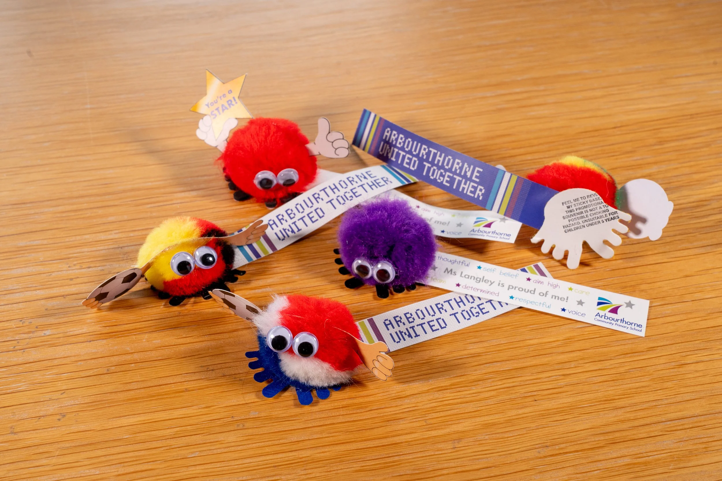 Colorful Pom-pom caterpillars on wooden surface with school-themed bracelets and messages.
