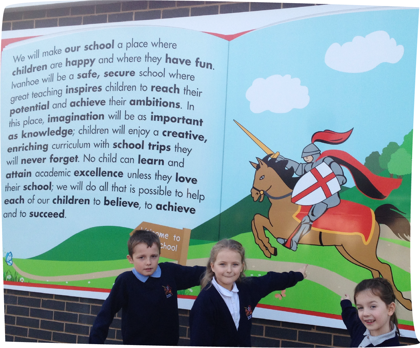 Children standing in front of a colorful school sign, pointing at an illustration of the Ivanhoe knight on a horse next to their school mission statement. The graphic has been designed to resemble a story book.