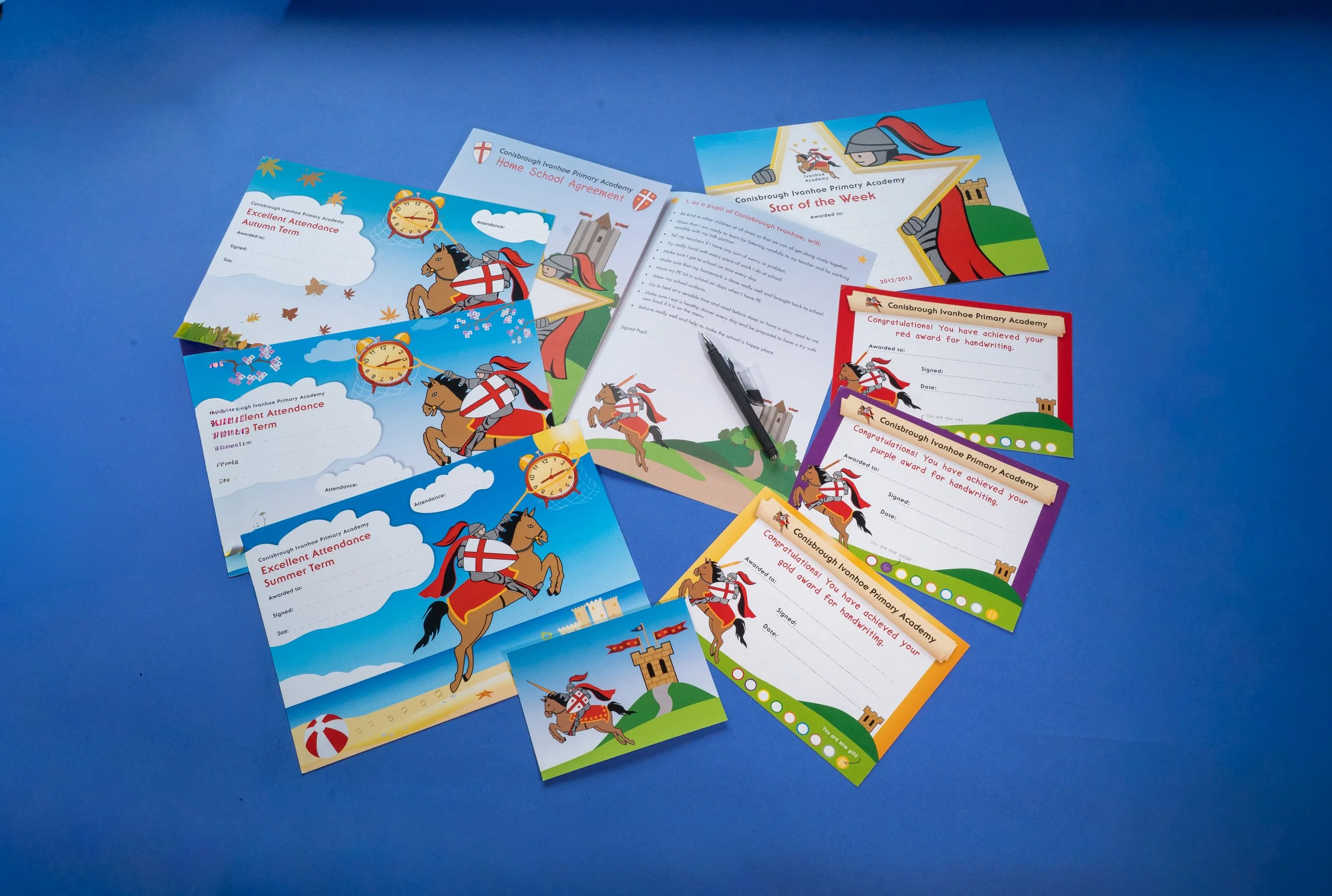 Colorful school newsletters and awards with illustrations of knights on horses, castles, and medieval-themed graphics, spread out on a blue surface.