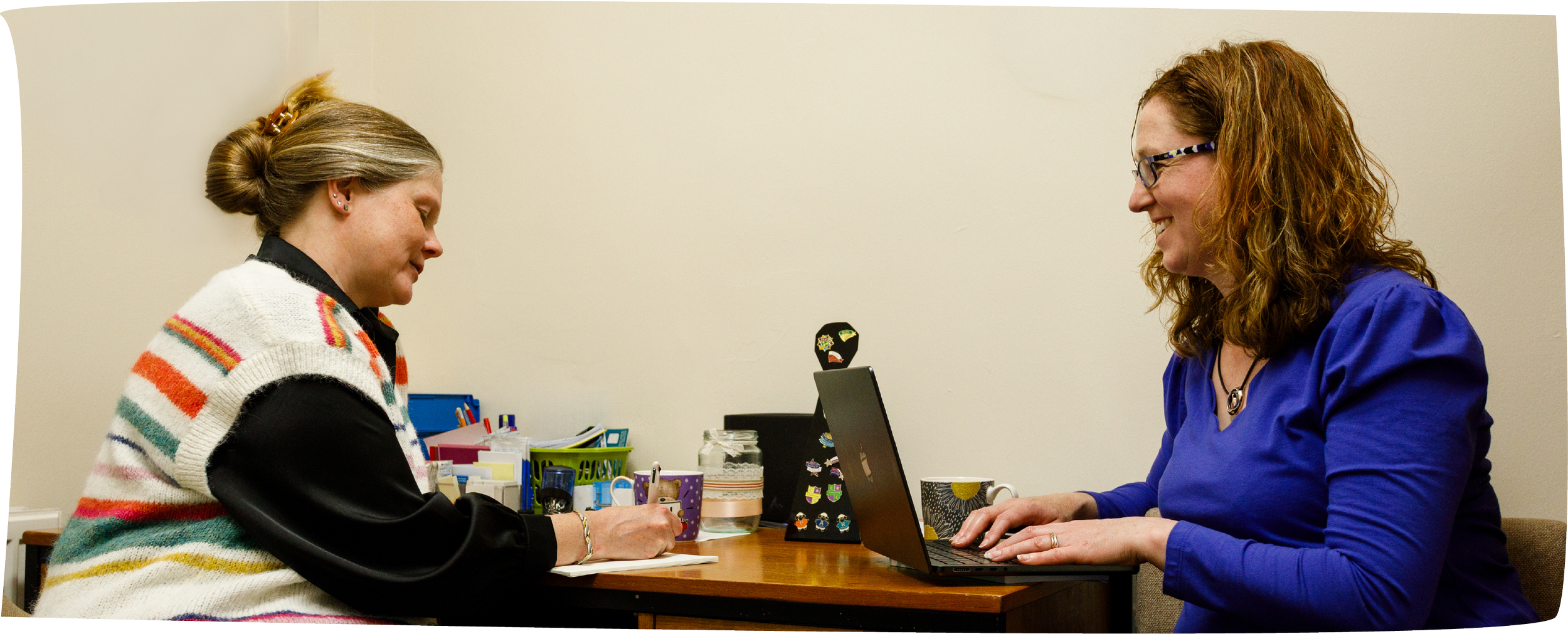Two women sitting at a table; one is writing in a notebook, the other is using a laptop; they appear to be having a conversation. The table has office supplies, mugs, and decorative items.
