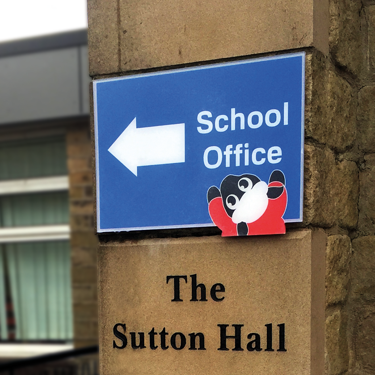 Blue sign with a white left arrow pointing to the school office, featuring a cartoon bear. Below, a stone plaque reads "The Sutton Hall."