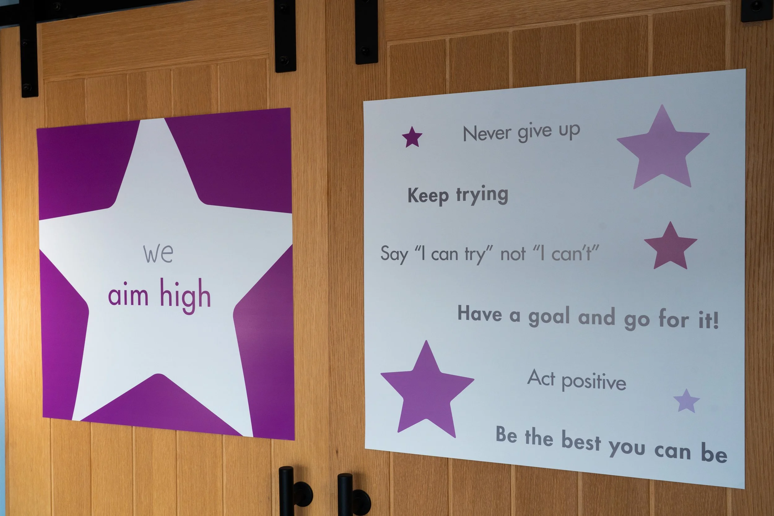 Two motivational posters on a wooden wall. The left poster has a purple star and the text "We aim high." The right poster has purple stars and the text "Never give up. Keep trying. Say 'I can try' not 'I can't'. Have a goal and go for it!"