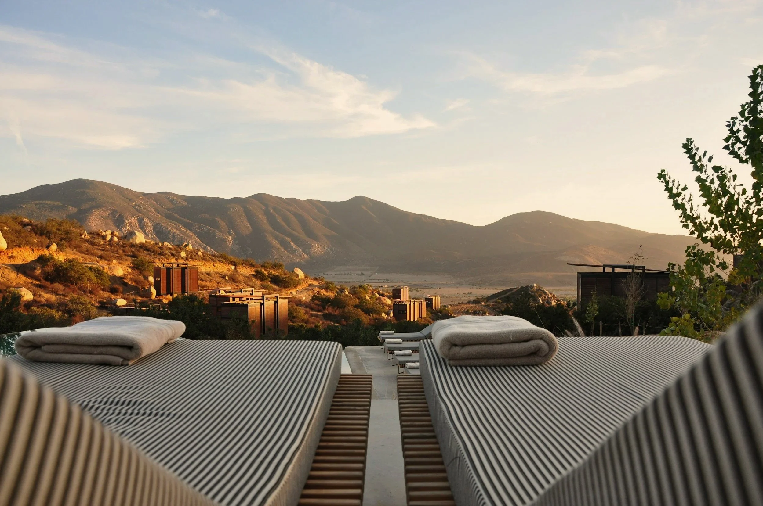 Exterior view of a mountain landscape with a clear sky, viewed from a terrace with striped lounge chairs and neatly folded towels.