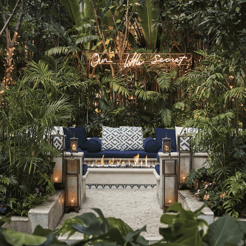 Luxurious indoor lounge surrounded by lush green plants with a neon sign reading 'Our Little Secret' above the seating area, featuring navy blue and white cushions and a modern fireplace.