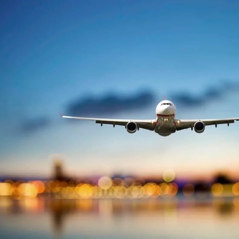 An airplane flying low over an airport runway at sunset with blurred city lights in the background.