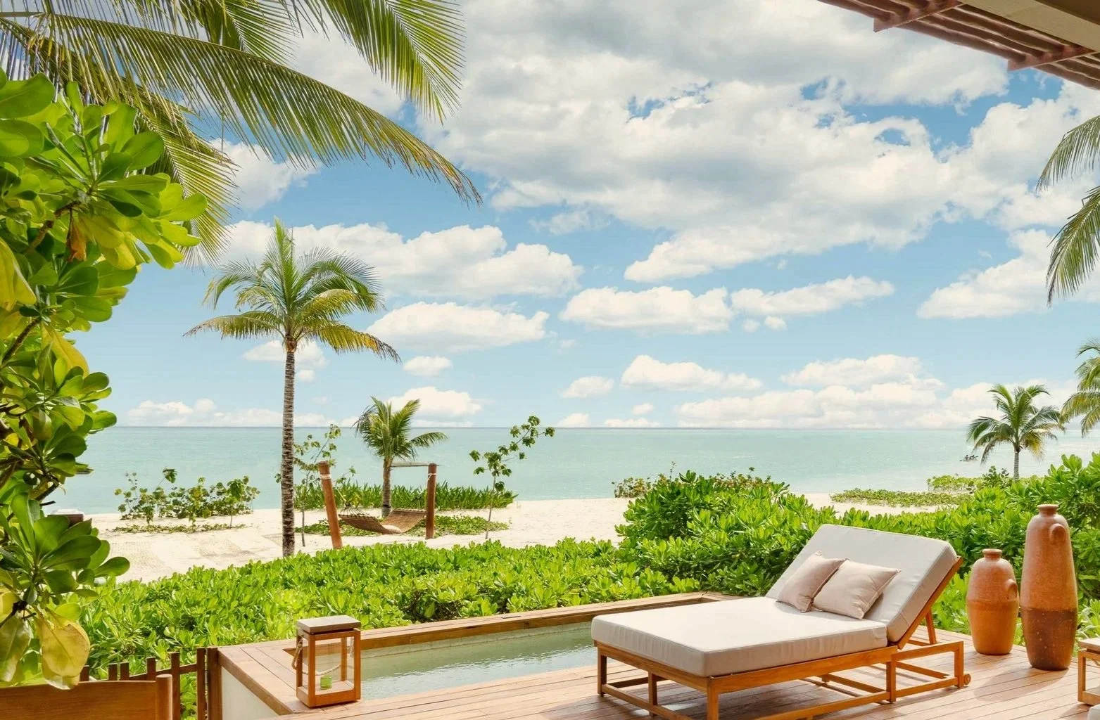 Rosewood Mayakoba, Mexico. Beach view from a patio with a lounge chair, large clay vases, surrounded by lush green plants, palm trees, and ocean in the background under a partly cloudy sky.
