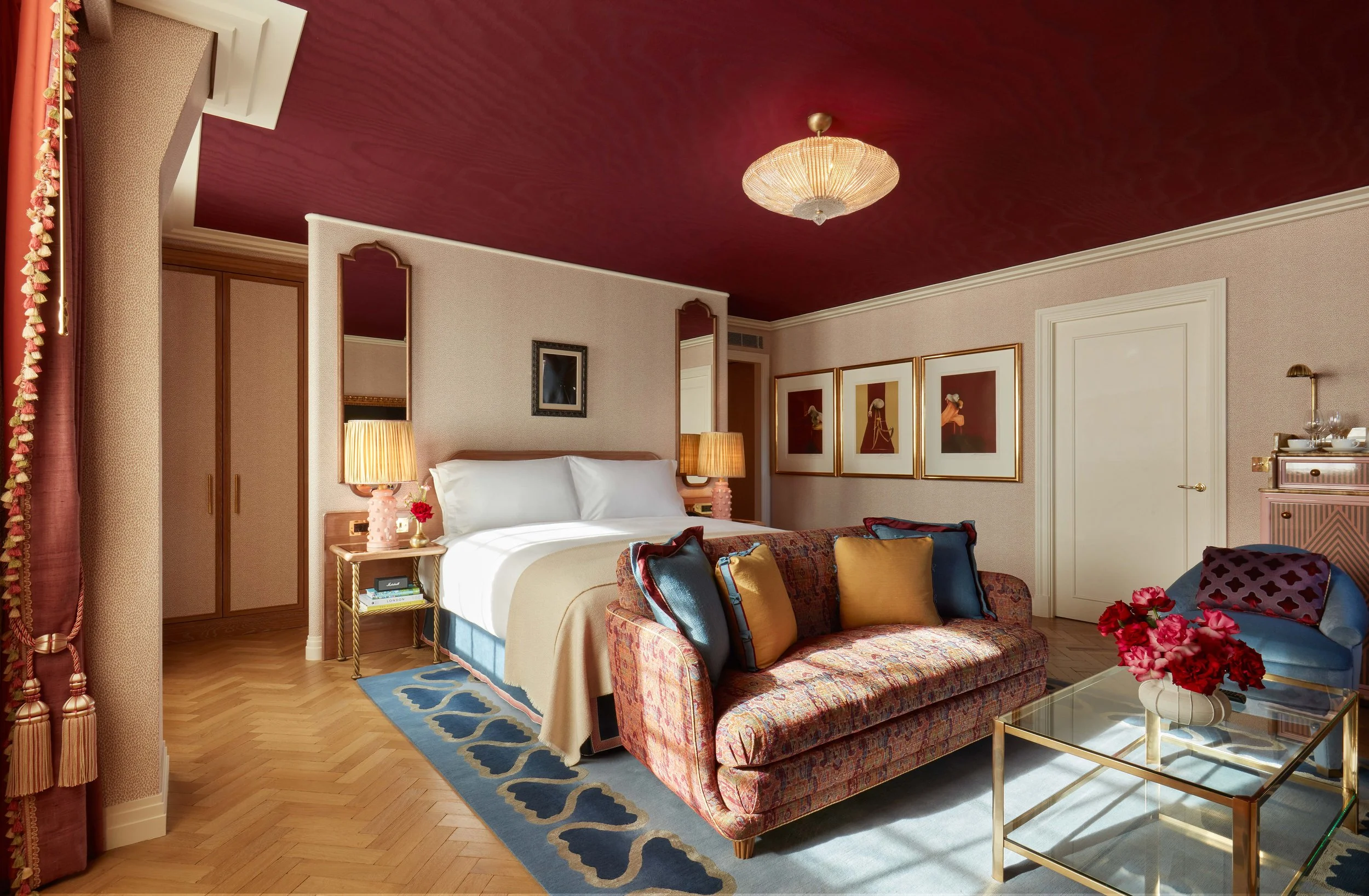 A luxurious hotel room with a bed, a floral sofa, and a glass coffee table. The room has a red ceiling, beige walls with framed artwork, and gold accents. There are two table lamps on bedside tables, and a closet with sliding doors.