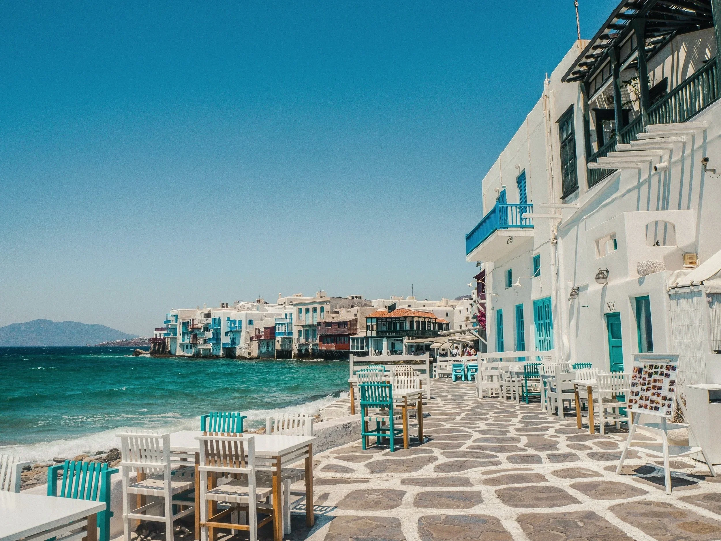 Coastal street in Greece with white buildings featuring blue doors and balconies, outdoor seating with turquoise and white chairs, and the sea with a clear sky.
