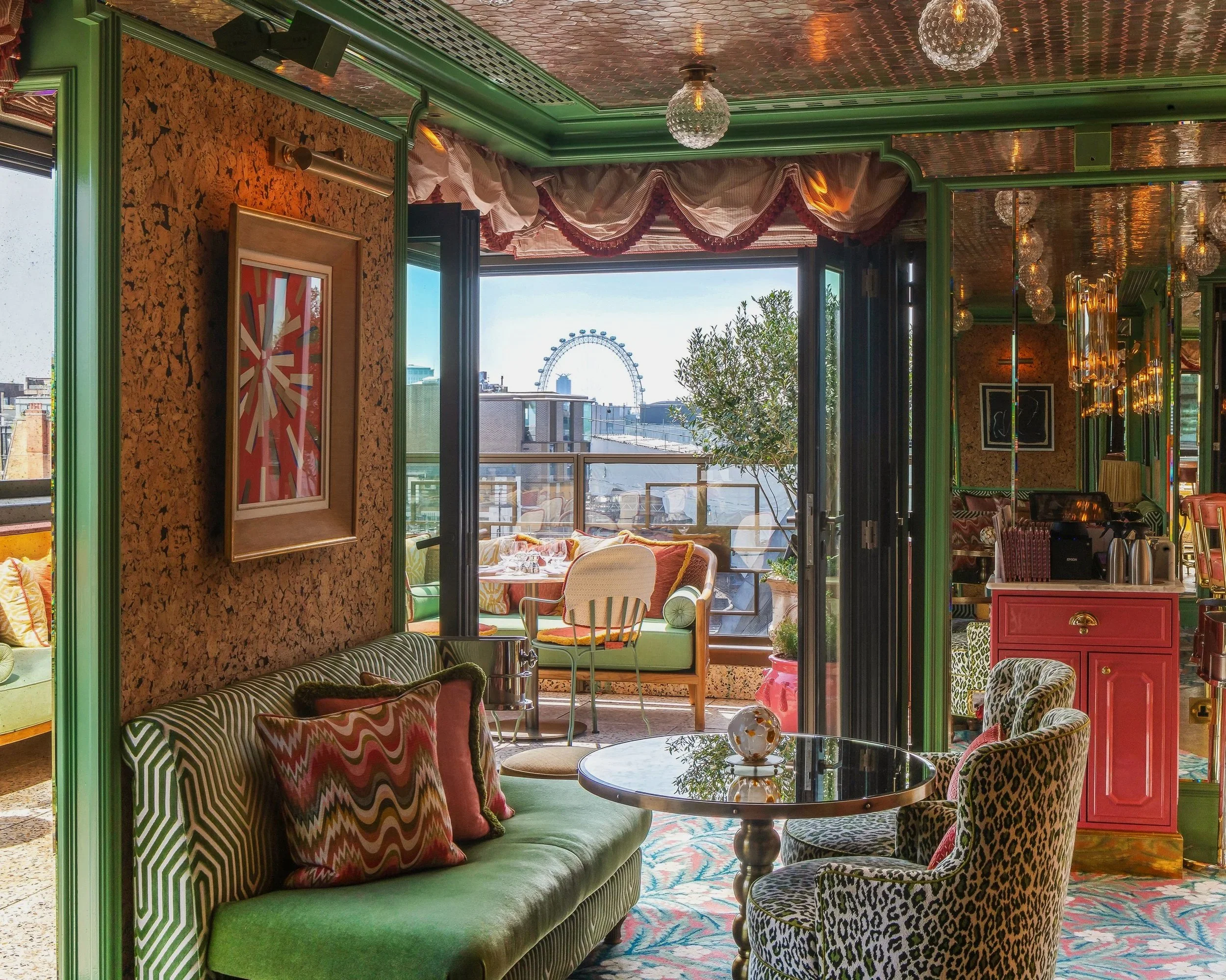 A vibrant living space with a view of the London Eye in the background. The room features colorful furniture including a green sofa with patterned pillows, a leopard print armchair, various decorative mirrors, and eclectic lighting fixtures. Large windows open to a balcony with outdoor furniture.