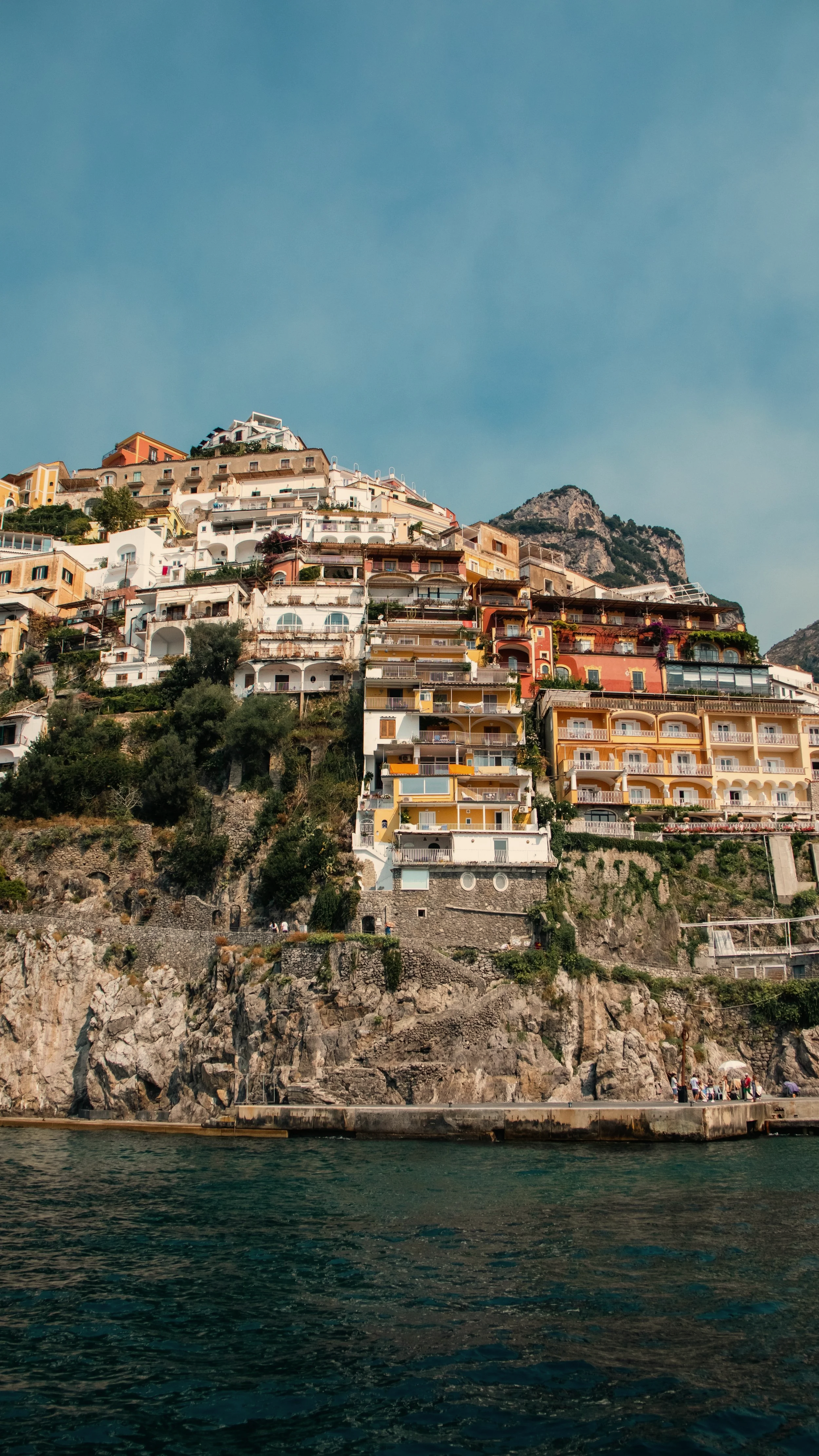 Positano, Italy. Colorful houses built into a rocky hillside overlooking the water, with a clear blue sky above.
