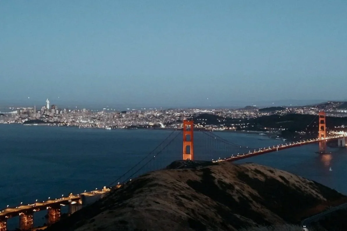 Night view of the Golden Gate Bridge with the San Francisco city skyline in the background, seen from a hillside, with water and a clear sky.