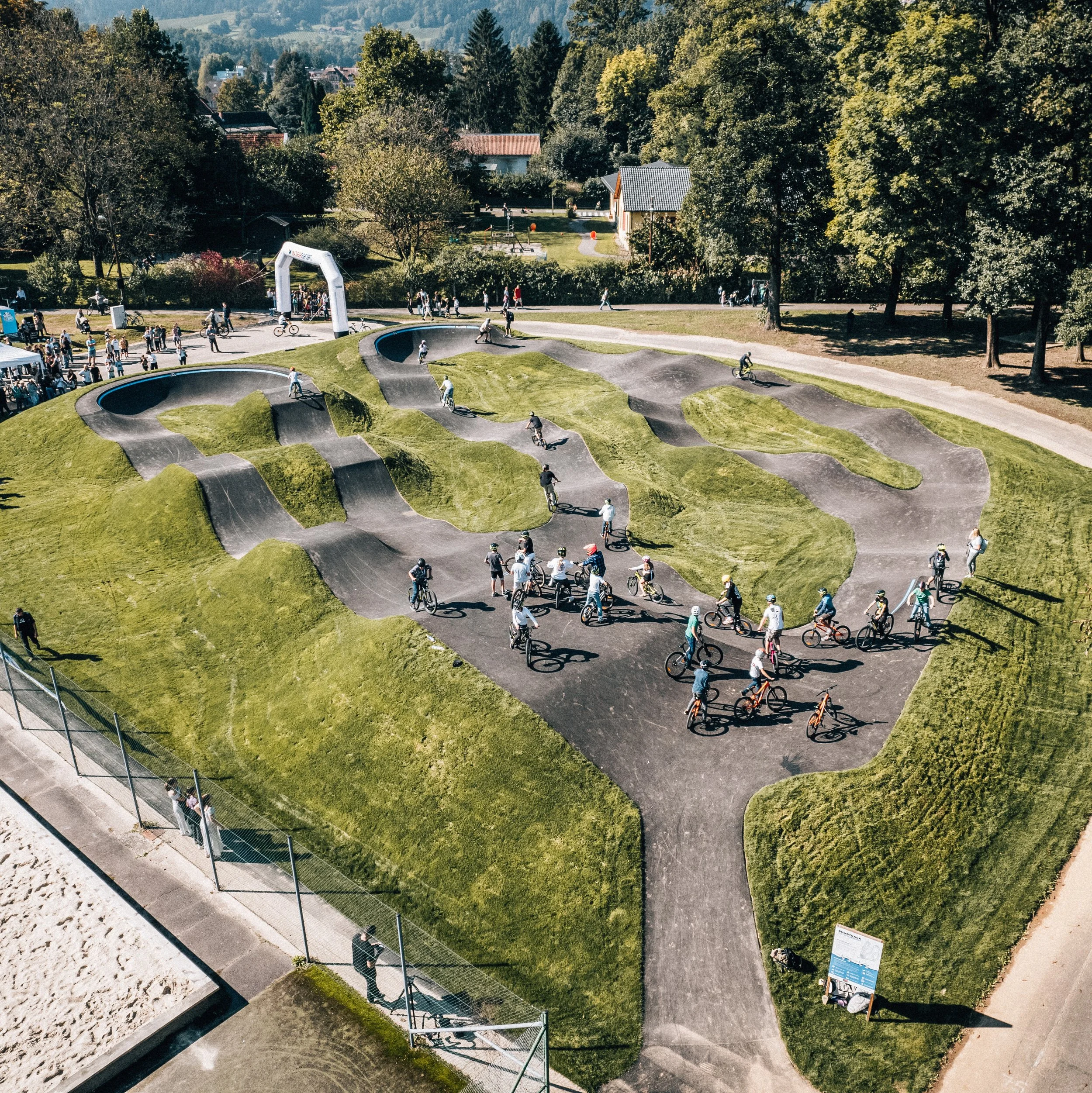 Eine Grünanlage mit einem Pumptrack für Alle, auf dem viele Menschen fahren und und einige im Startbereich stehen.