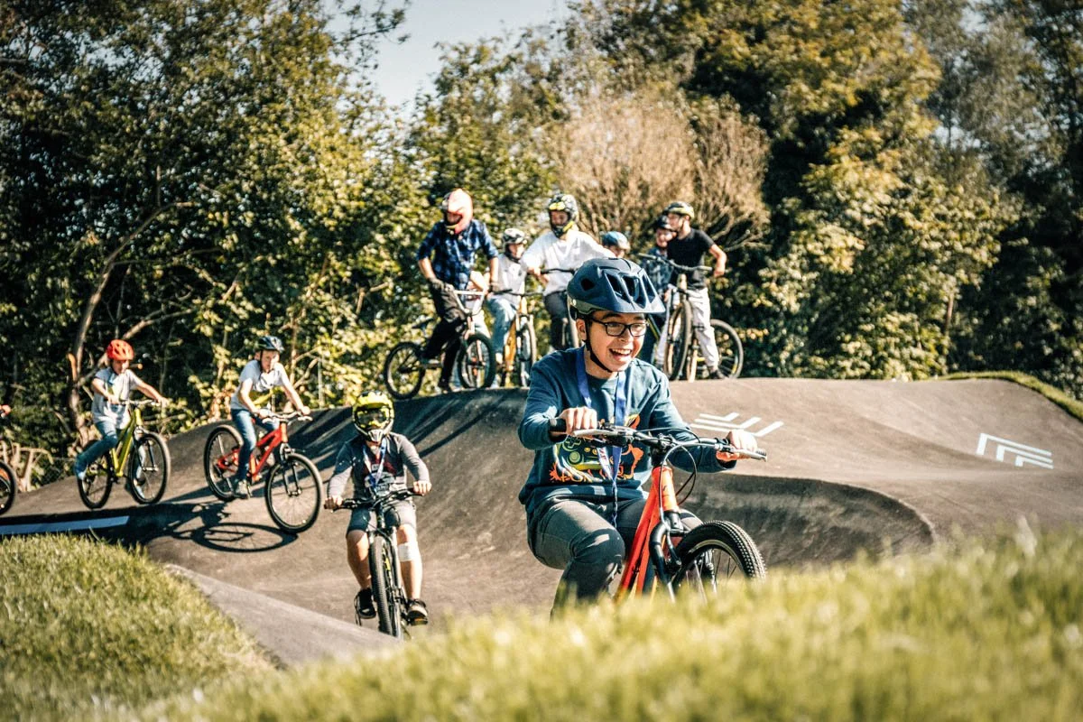 Kinder und Jugendliche beim Fahren auf einem Pumptrack im Freien an einem sonnigen Tag, umgeben von Bäumen.