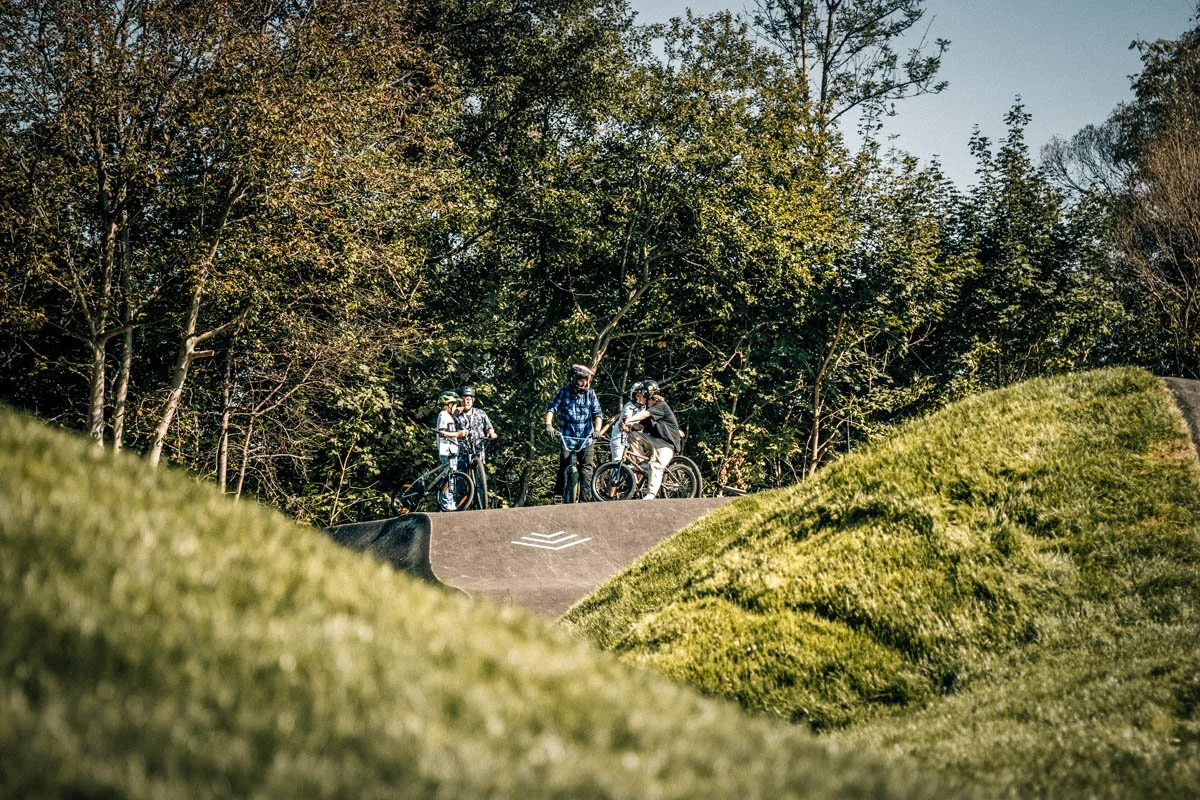 Gruppe von Jugendlichen mit Fahrrädern auf einem Pumptrack im Grünen, umgeben von Bäumen.