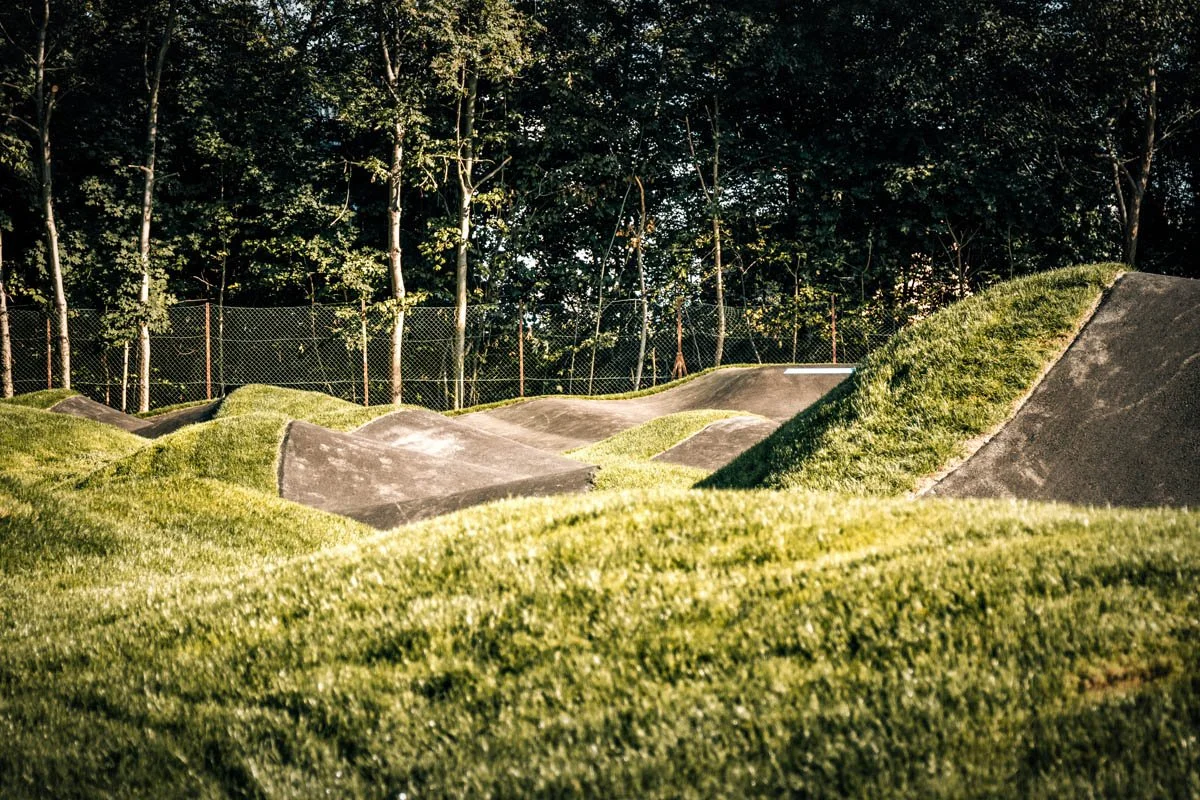 Ein Pumptrack mit Asphaltflächen, umgeben von grünem Gras und Bäumen im Hintergrund.