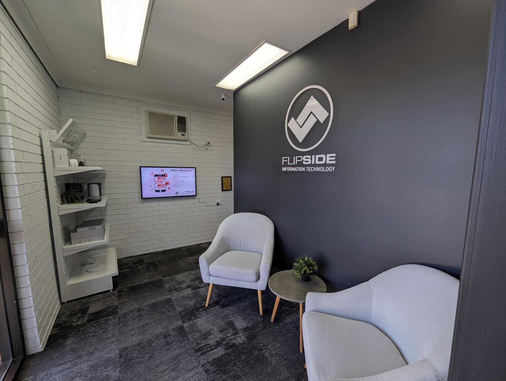 Office reception area with a gray wall displaying the company logo 'FLIPSIDE INFORMATION TECHNOLOGY', white chairs, a small round table with a potted plant, a wall-mounted monitor, white brick walls, and an air conditioning unit.