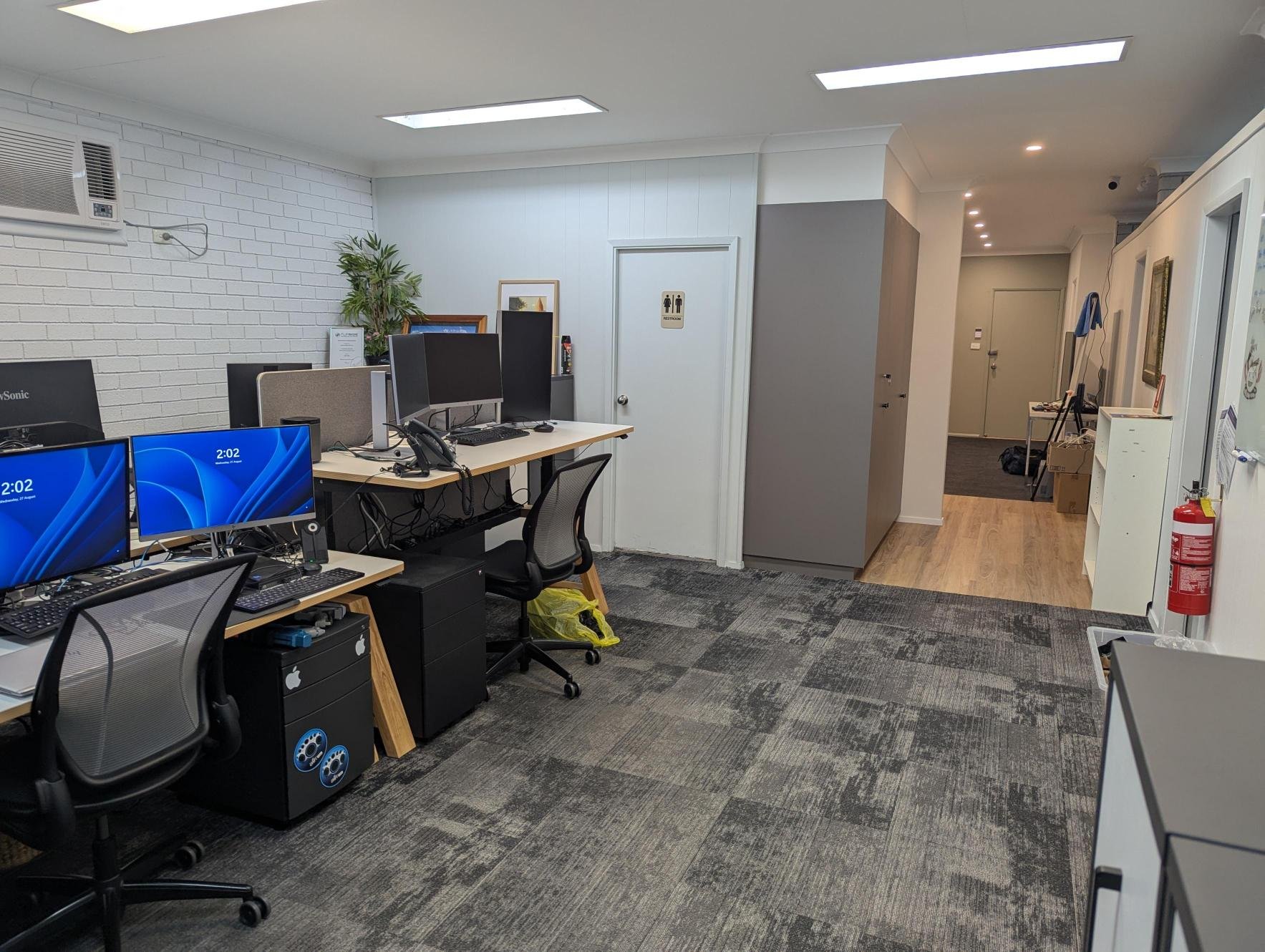 Office workspace with desks, computers, and chairs, a small potted plant, a framed picture, and a door with restroom sign, beige, white, and gray color scheme, fluorescent lighting, carpeted floor.