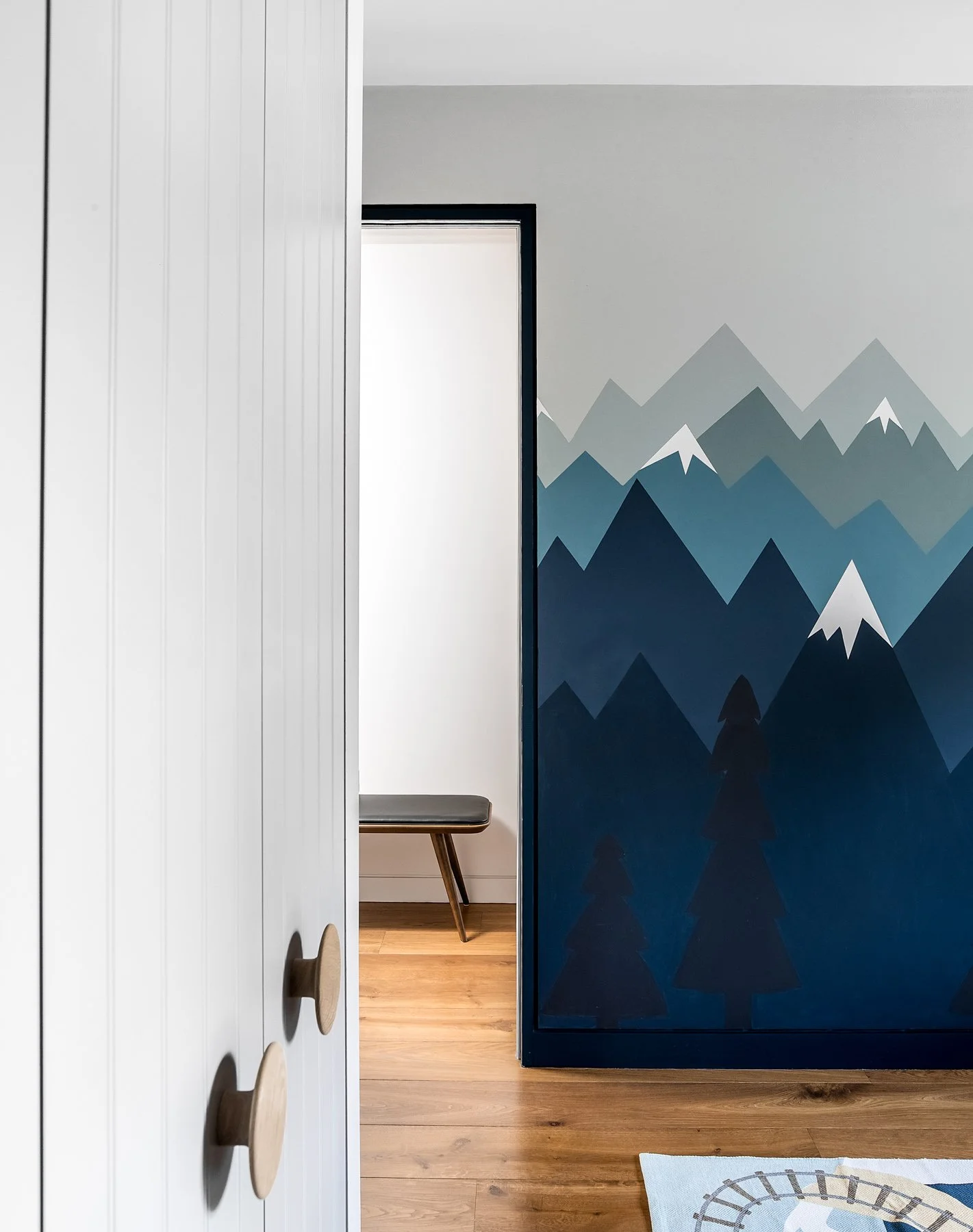 Interior of a modern room with a white wardrobe featuring circular handles on the left, a partial view of a bench with wooden legs and a gray cushion in the middle, and a blue wall with mountain and forest mural on the right. The room has wooden flooring and part of a colorful patterned rug is visible at the bottom.