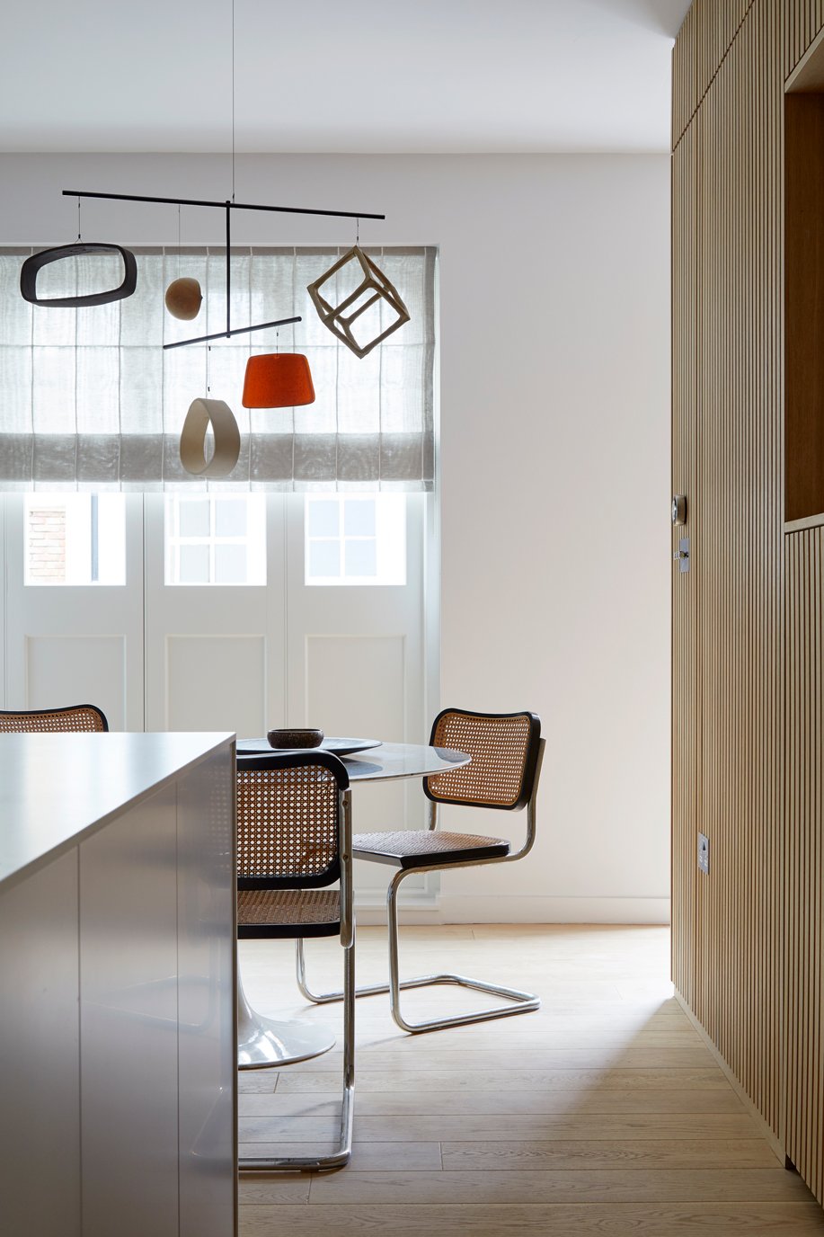 Modern dining area with a white table, two cane-back chairs, and a unique hanging light fixture with geometric and abstract shapes.
