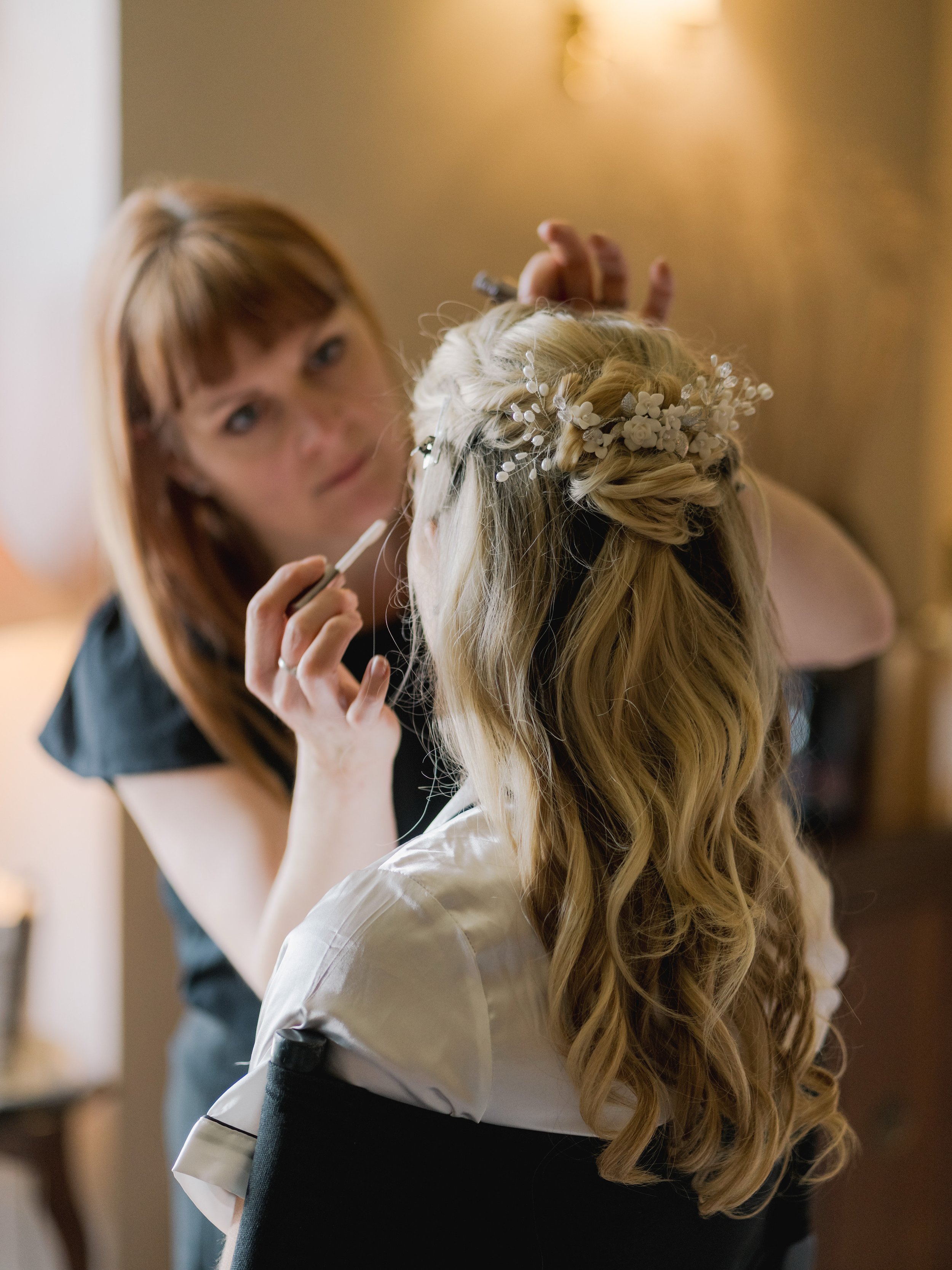 A woman with long, curly blonde hair wearing a decorative headpiece, possibly a bridal or special occasion accessory. Another woman is in the background, applying makeup or lipstick to the woman with the hairpiece.
