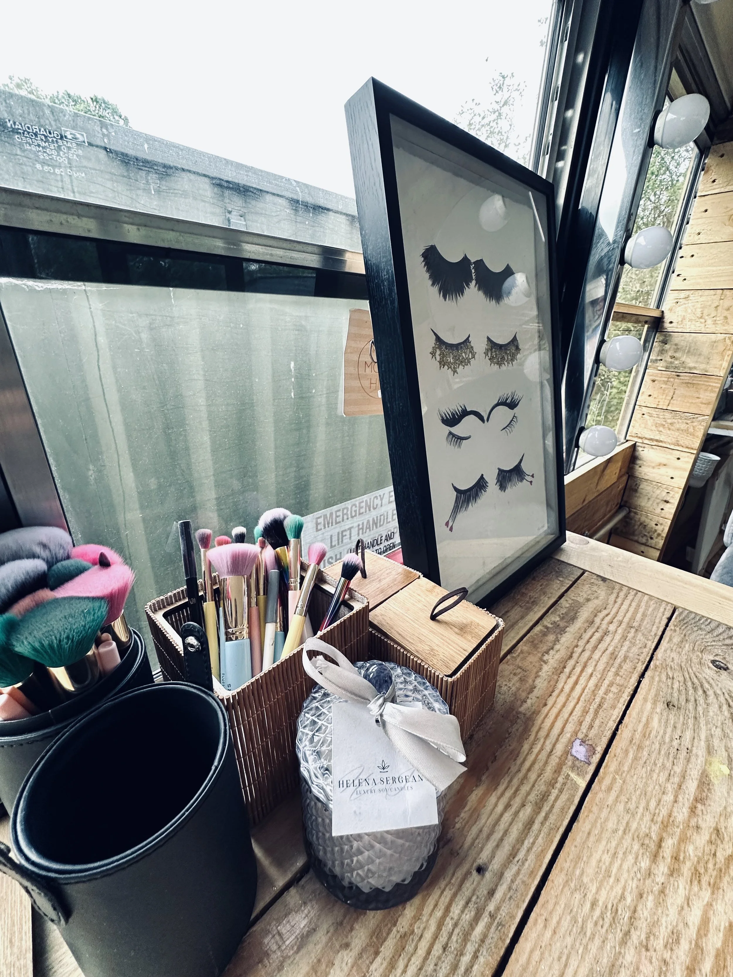 A wooden table holding makeup brushes, black containers, a paper bag with a bow, and a framed display of false eyelashes, with a window and wooden wall in the background.