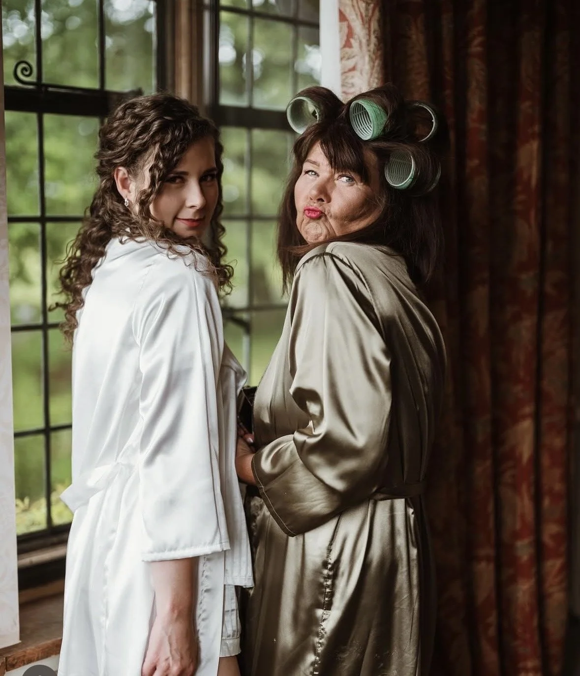 A young woman with curly hair in satin pajamas standing next to an older woman with satin robe and hair rollers in a room with large windows and red curtains, both making playful expressions.