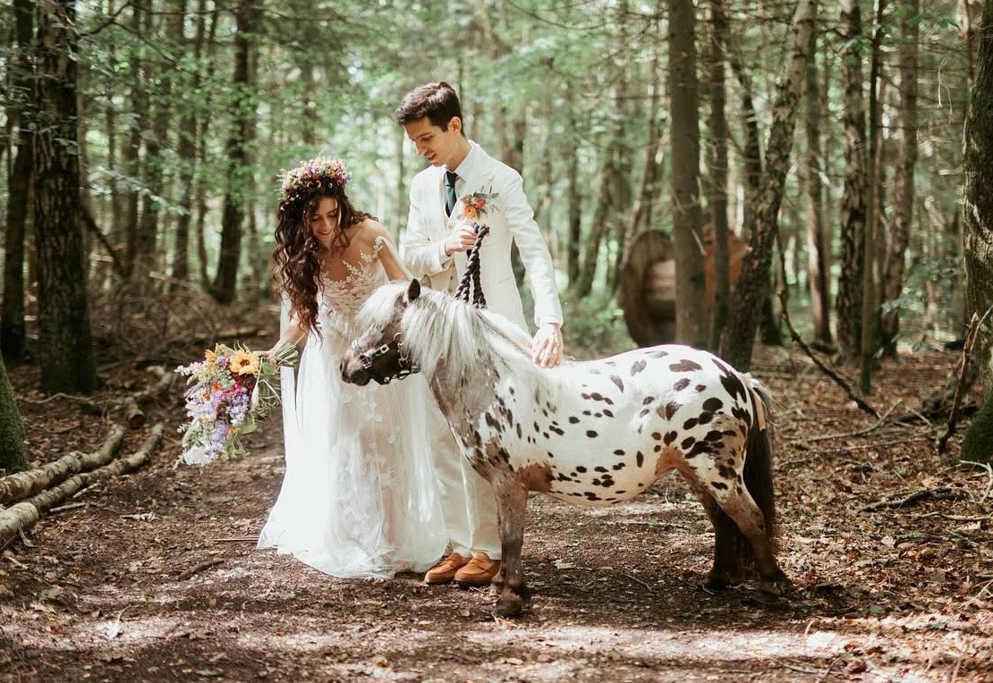 A newlywed couple in wedding attire stands in a forest, with the bride holding a bouquet of flowers and the groom holding the reins of a small, spotted horse.