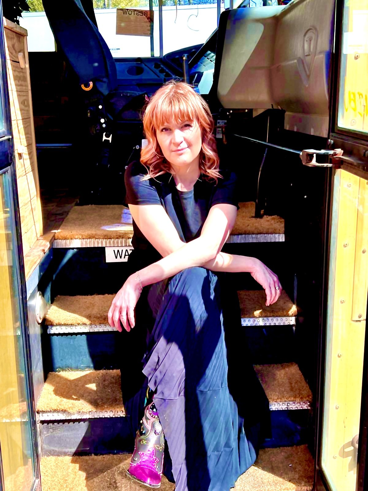 Woman with reddish hair sitting on stairs outside a yellow booth, sunlight on her face, wearing a black top, blue pants, and colorful shoes.