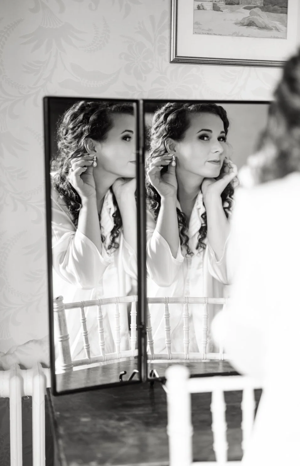 A woman with curly hair is looking at her reflection in a mirror, touching her earring, while she is getting ready. The scene is black and white, with a framed picture on the wall and a wooden chair in front of the mirror.