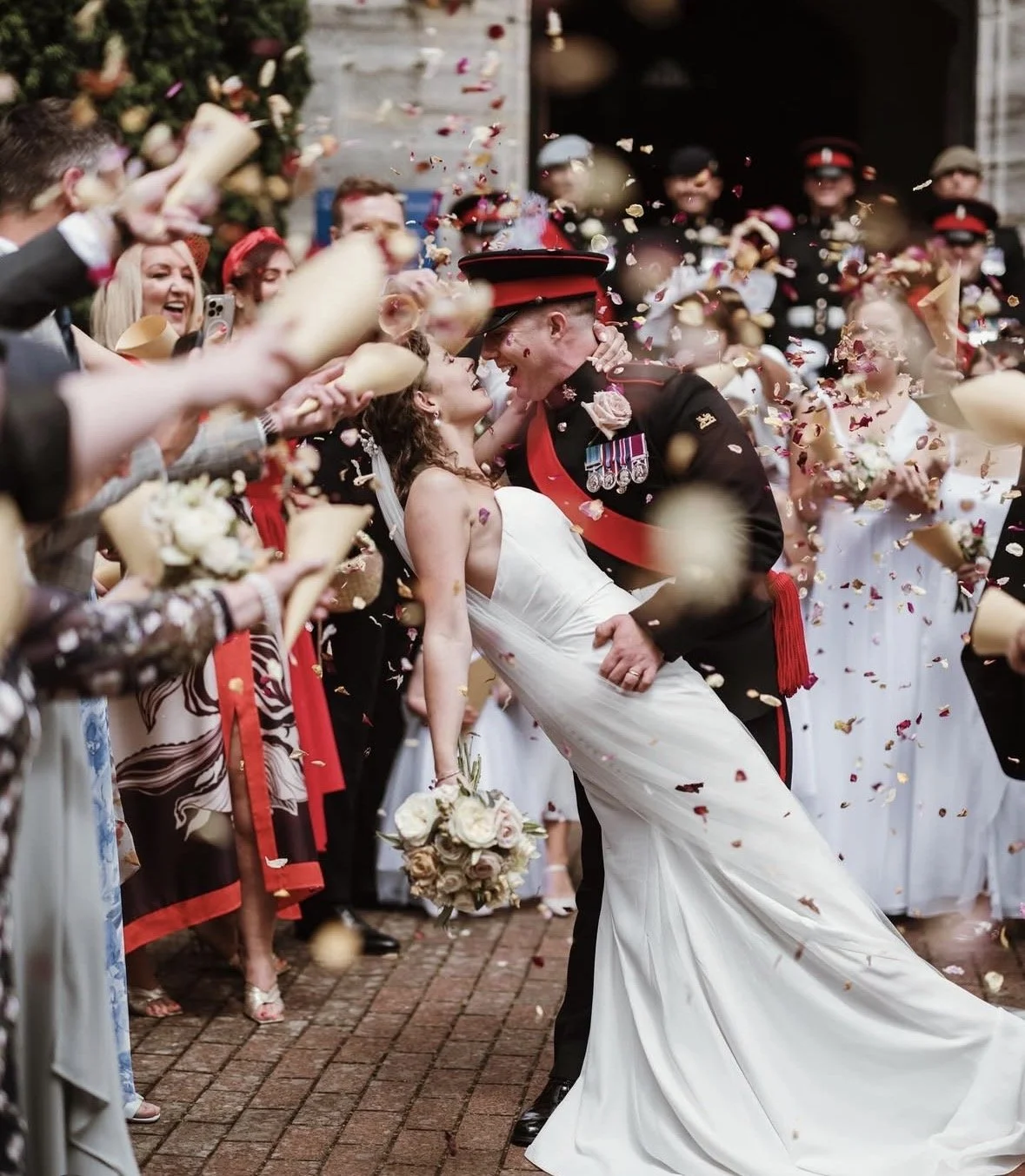 A bride and a soldier in uniform share a romantic moment during a wedding celebration, surrounded by guests throwing confetti and celebrating.