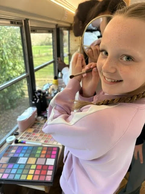 Young girl smiling and applying makeup in front of a mirror inside a bus or van, with a makeup palette on a table nearby and a scenic outdoor view through the window.