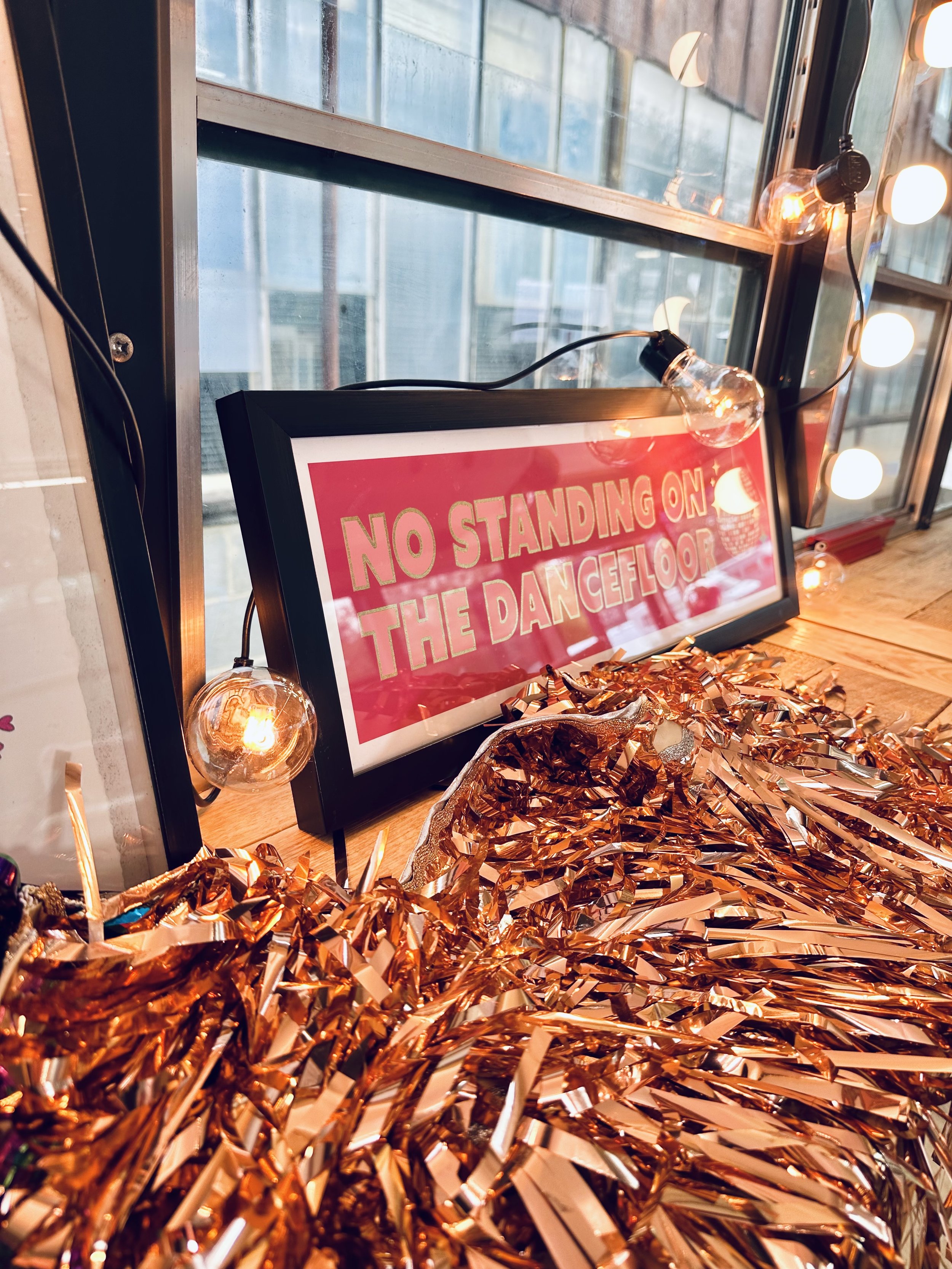 A sign reading 'No Standing on the Dance Floor' is displayed behind a string of warm yellow string lights, with metallic rose gold tinsel on a table in the foreground.