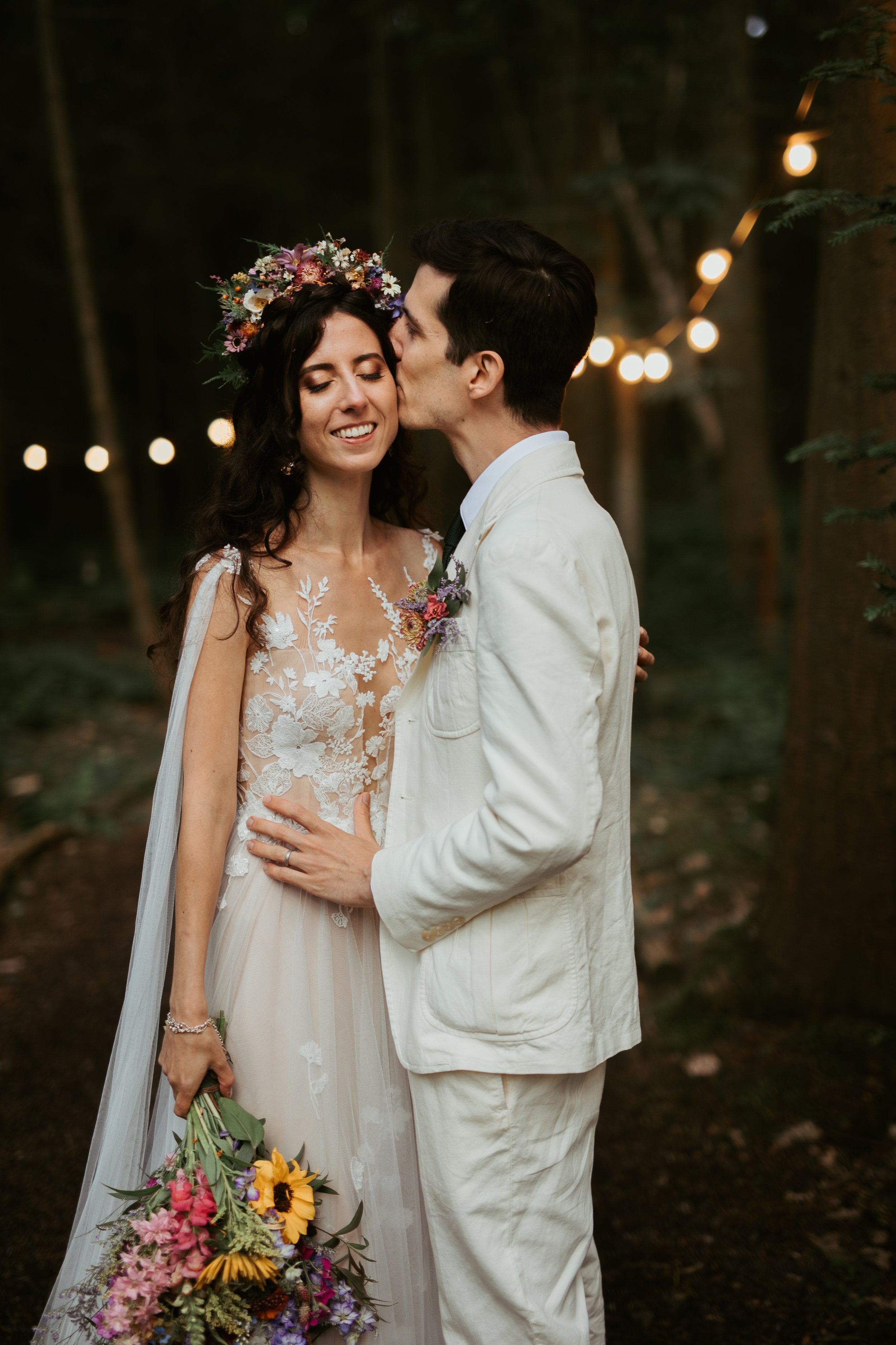 A bride and groom sharing a kiss in a wooded outdoor setting at night, with string lights in the background. The bride wears a floral crown and a lace wedding dress, holding a large bouquet of colorful flowers, and has a joyful expression with her ey