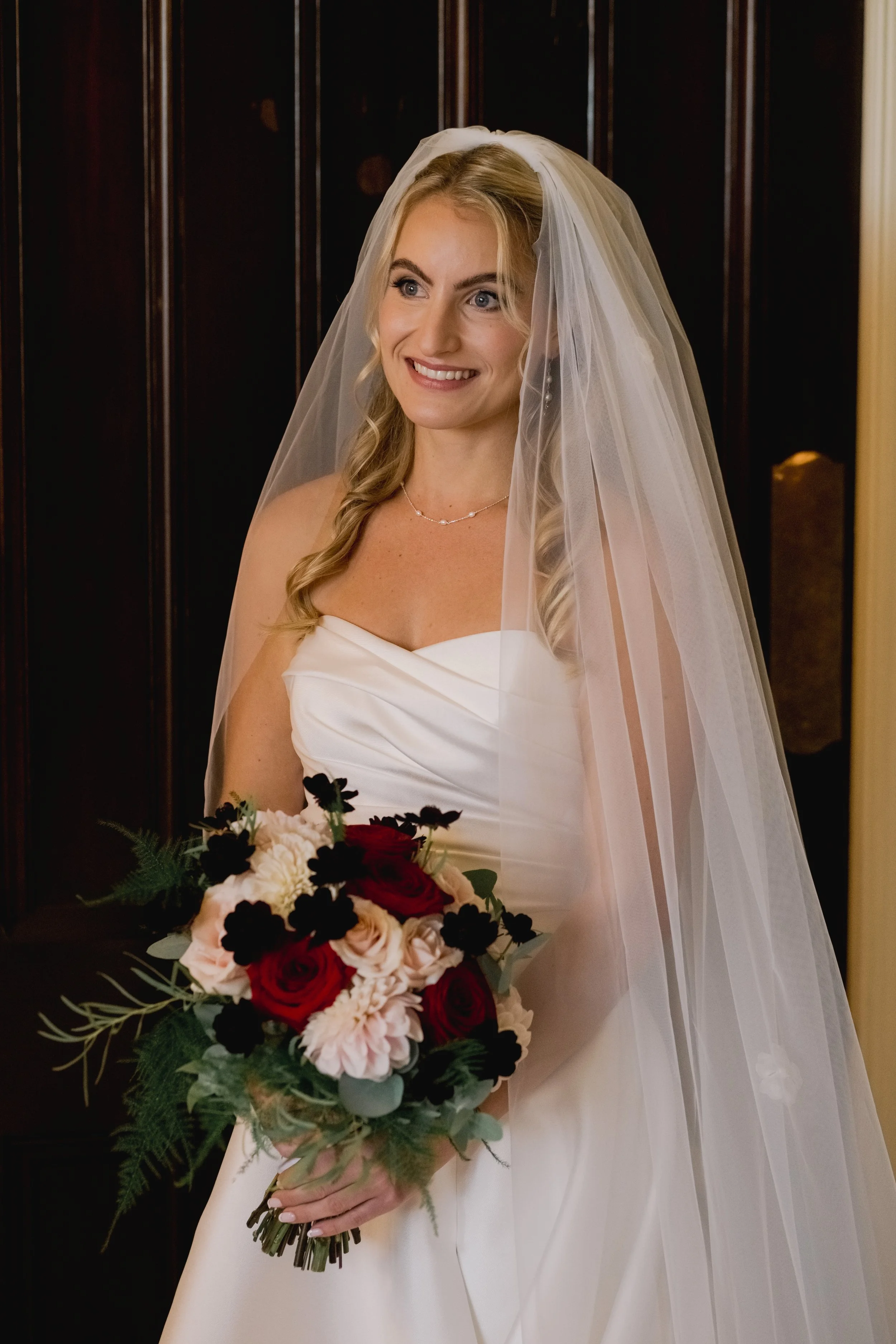 A smiling bride wearing a white strapless wedding gown, veil, and jewelry, holding a bouquet of red, pink, white flowers, with a dark wooden background.