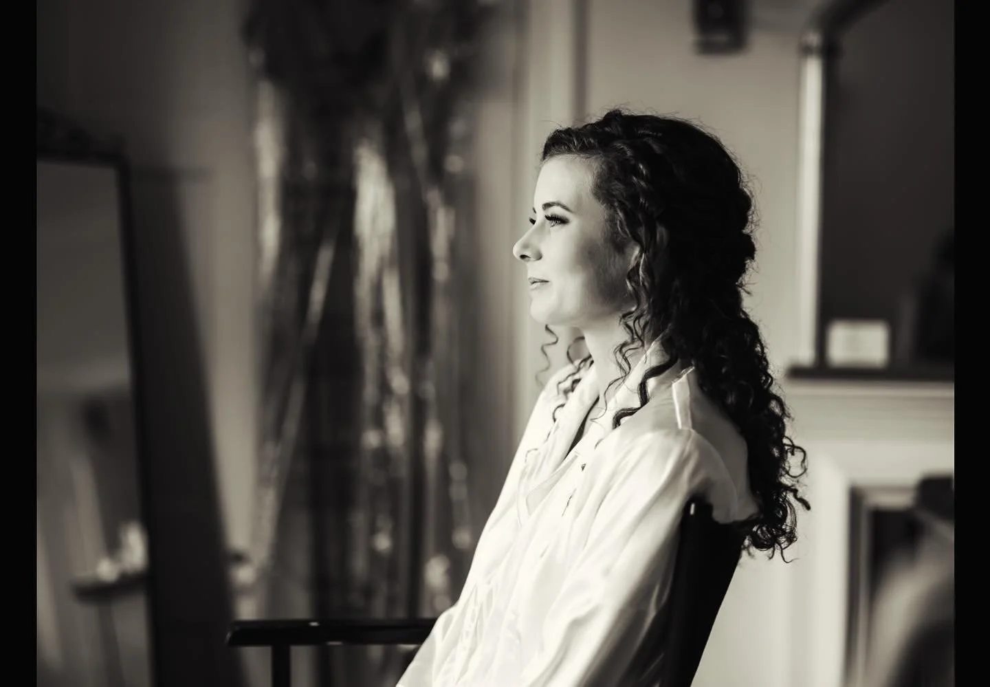 A woman with curly hair sitting in profile, looking out a window, in a softly lit room, black and white photograph.