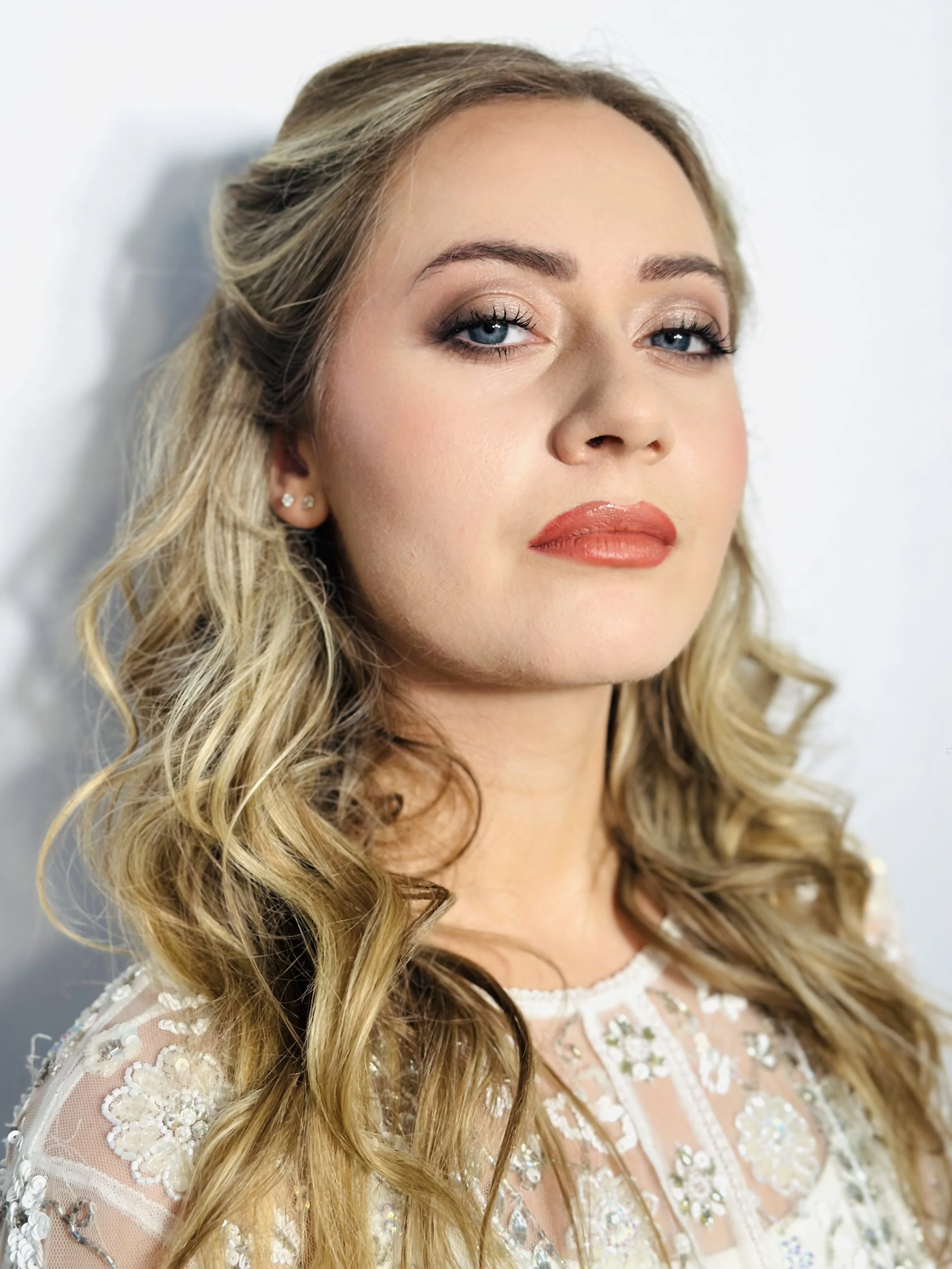 Close-up of a woman with blonde, curly hair and blue eyes, wearing makeup with gold eyeshadow and coral lipstick, in a white lace dress with intricate floral embroidery.