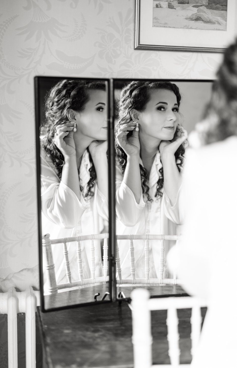 A woman with curly hair is looking into a mirror, adjusting an earring, with her reflection visible in a double-sided mirror. The scene is in black and white inside a room with patterned wallpaper and a framed picture on the wall.
