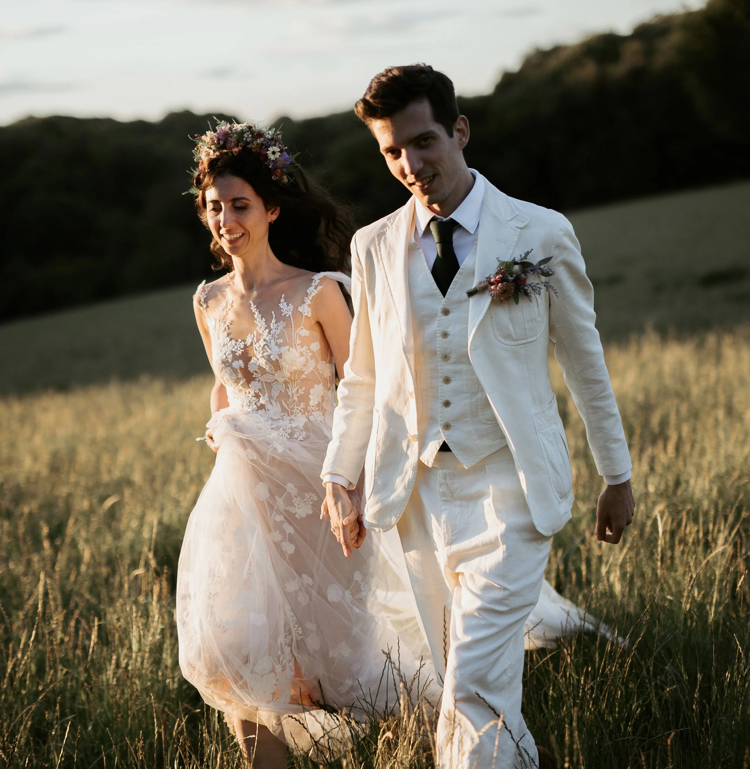 A couple dressed in wedding attire holding hands while walking through a grassy outdoor field at sunset.