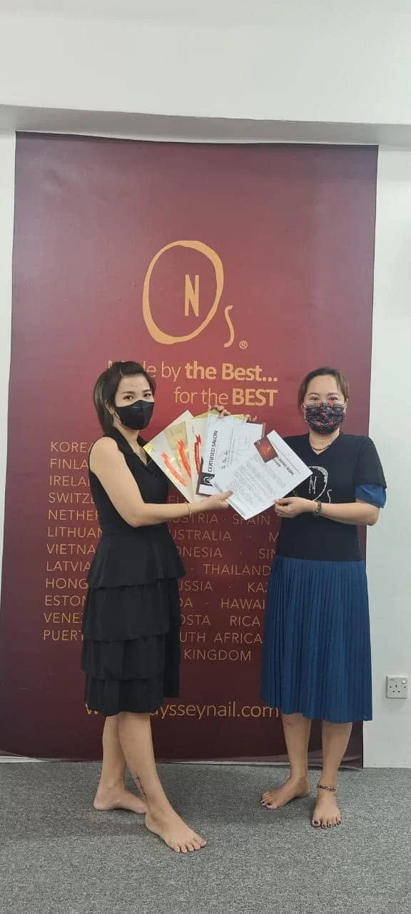 Two women standing indoors, holding certificates, and wearing masks. They are barefoot and standing in front of a maroon backdrop with gold writing and a large logo at the top.