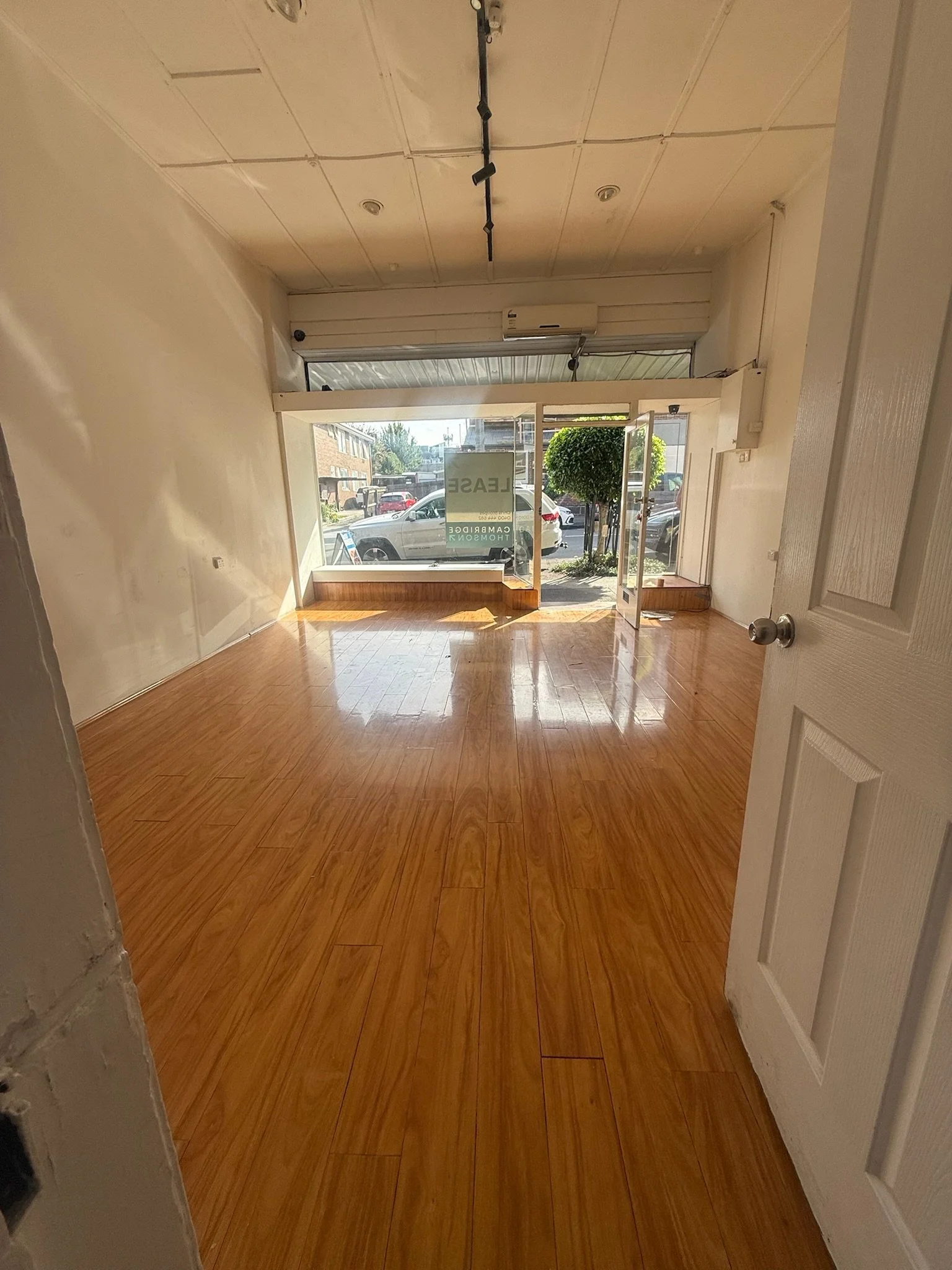 Empty retail space with large glass storefront, wood flooring, and an A/C unit on the back wall. A 'For Lease' sign is visible outside in the window.