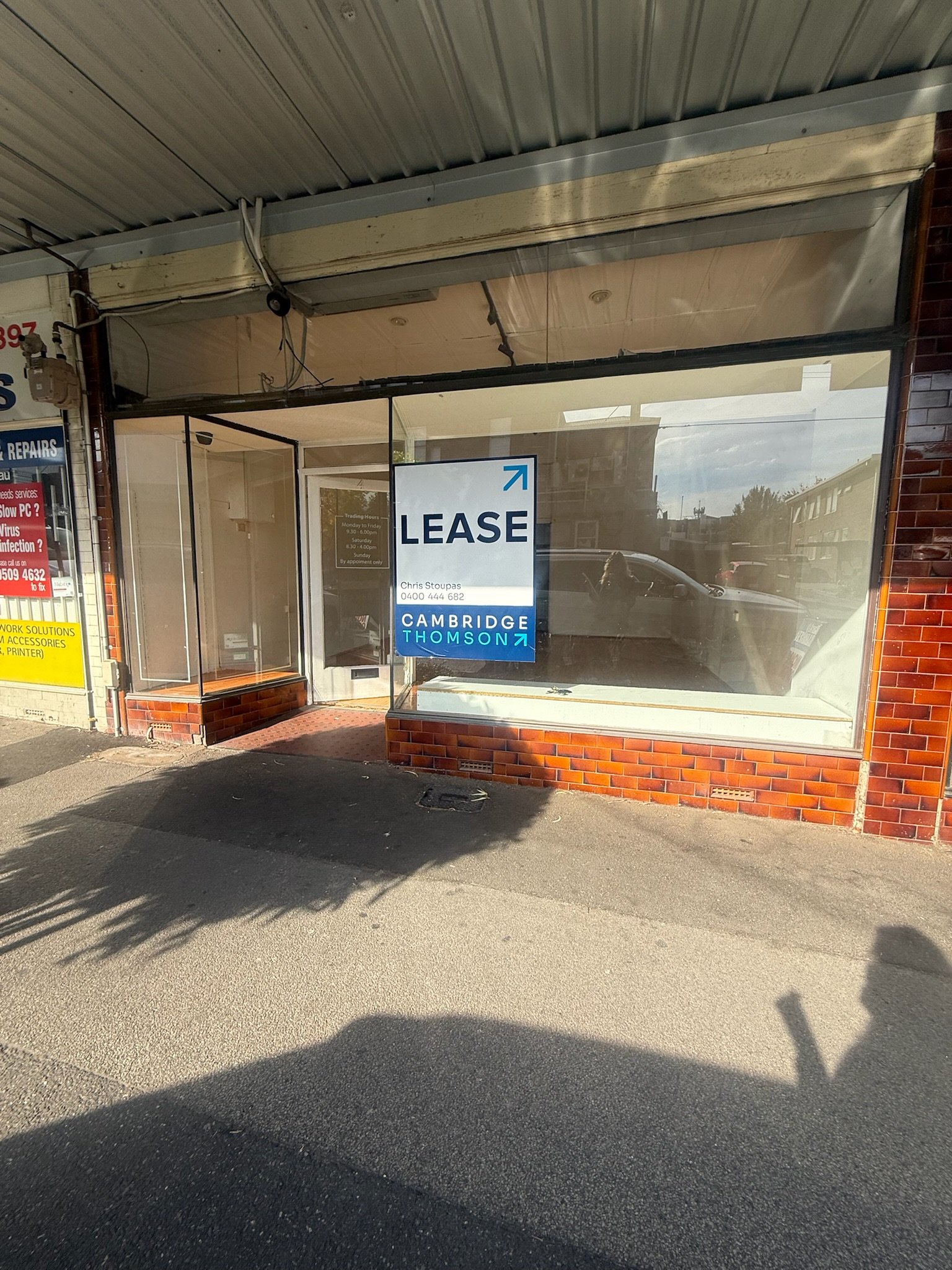 Empty storefront with large window and lease sign, brick facade, sidewalk, parked cars reflected in the glass, and shadows cast on the ground.