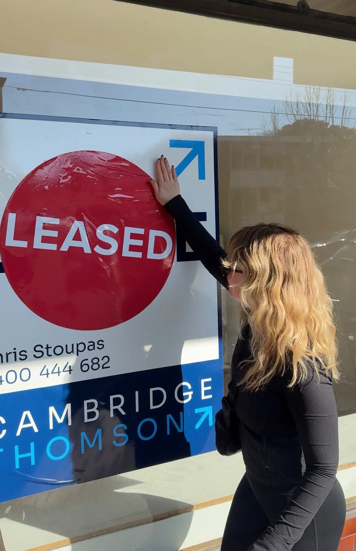 Woman peeling off a red 'Leased' sticker from a real estate sign outside a building.