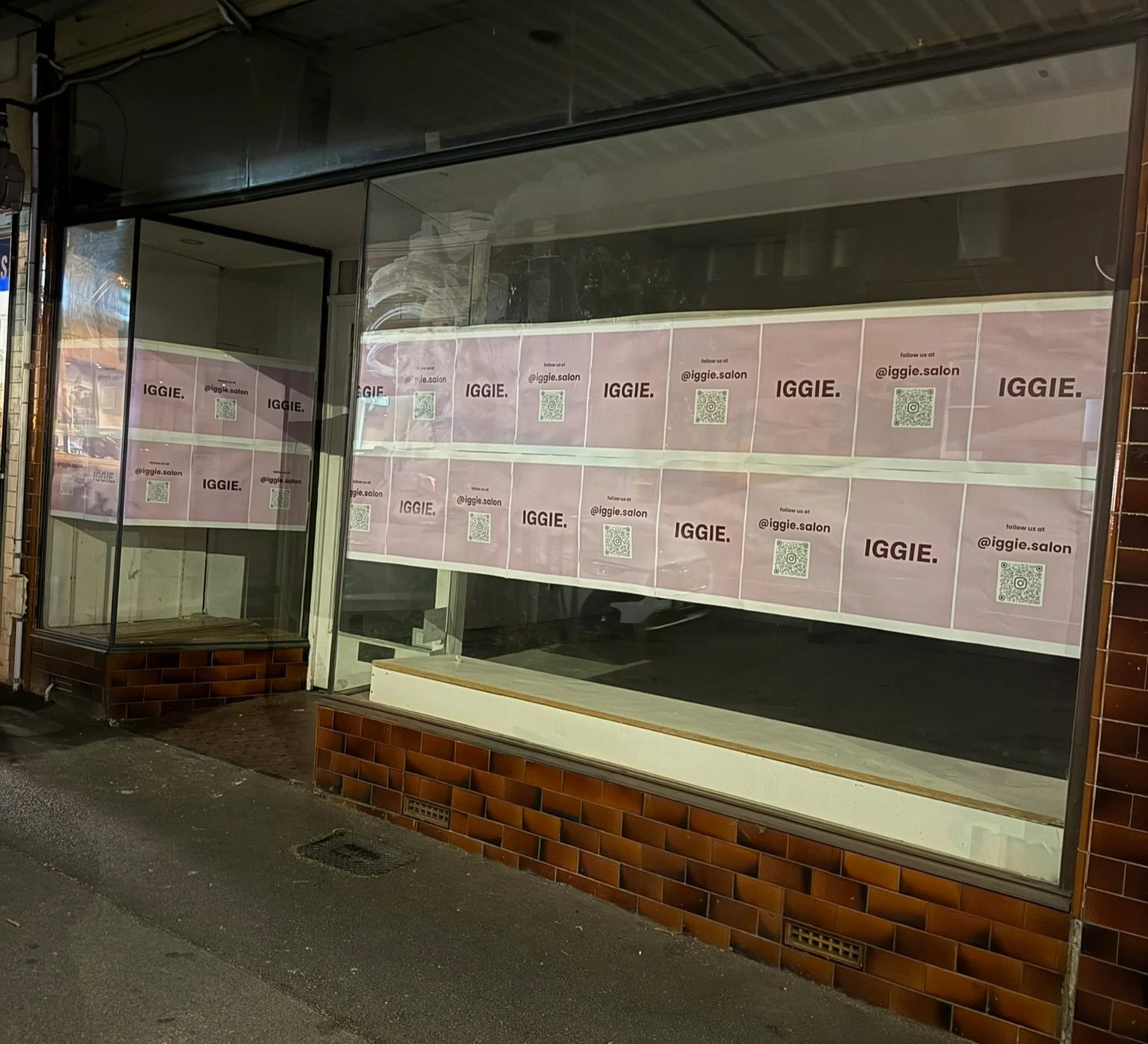 Empty storefront window with pink posters displaying the word IGGIE, a QR code, and social media handle @iggie.salon.